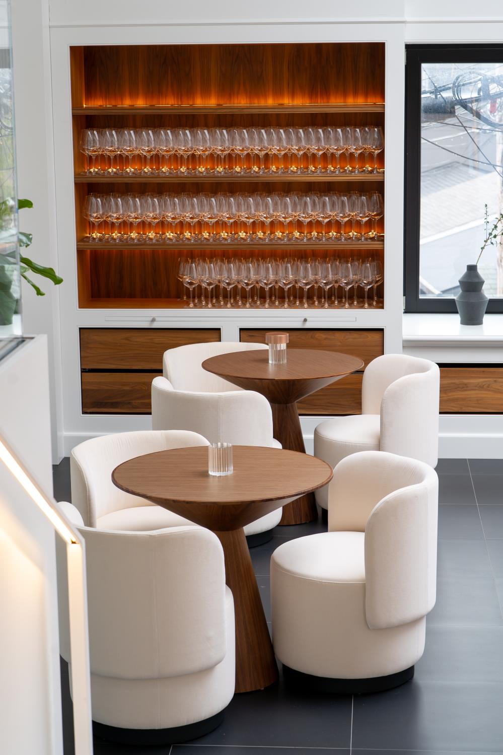 Modern dining area with two round wooden tables and six white upholstered chairs in front of a wooden cabinet filled with neatly arranged wine glasses.