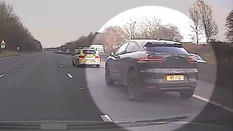 Dashcam view of a dark grey Jaguar SUV with visible rear license plate driving on a multi-lane motorway, highlighted by a circular vignette, with a marked police car ahead and trees along the roadside.