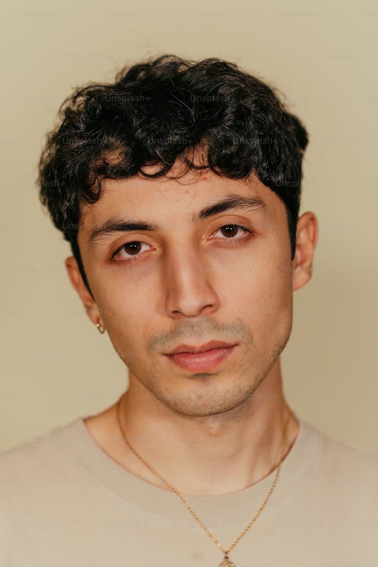 Young man with curly black hair wearing a beige shirt and gold necklace against a plain beige background.
