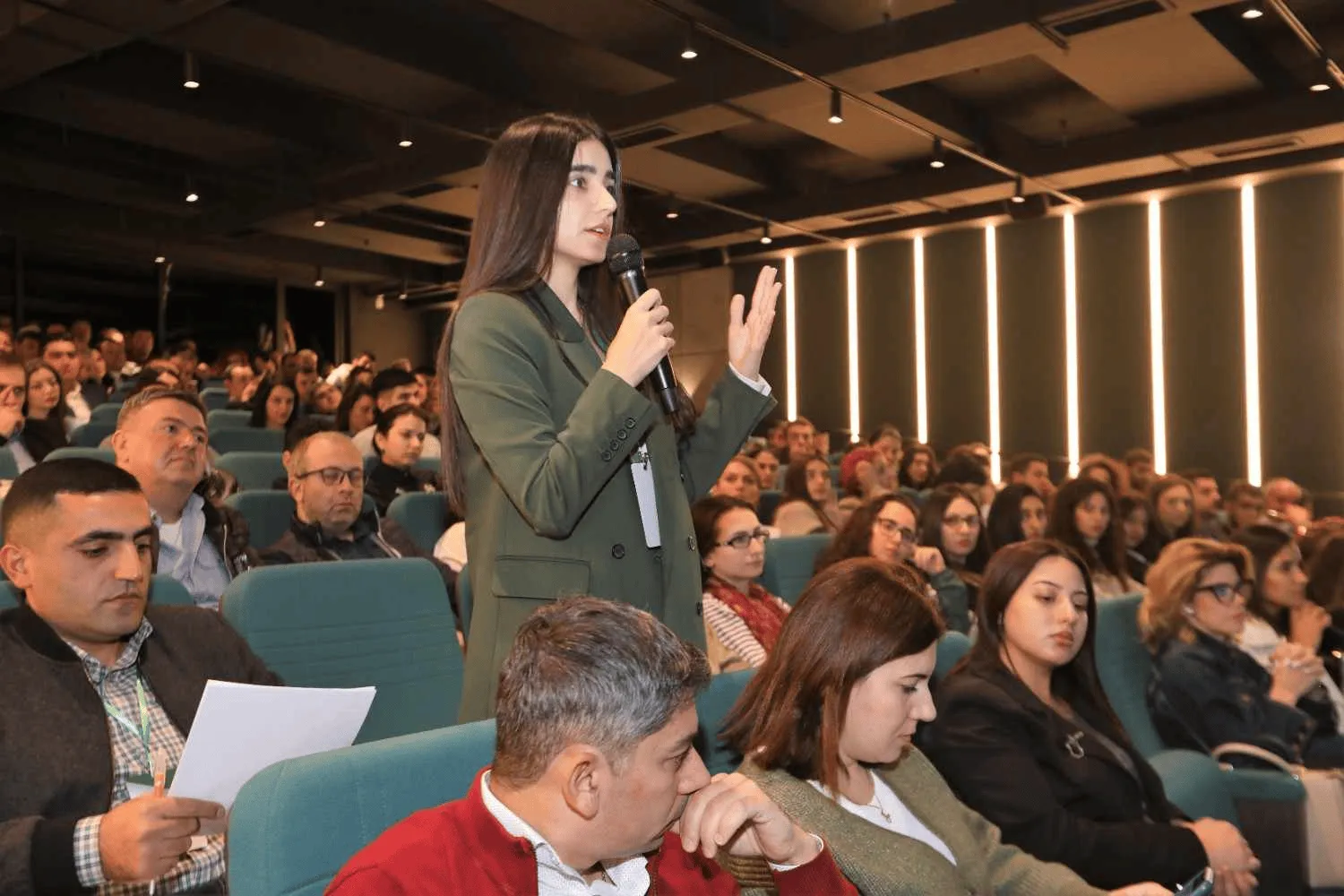 A woman in a green blazer stands in a conference room holding a microphone and speaking to an audience seated in blue chairs, with many listeners attentively watching her.