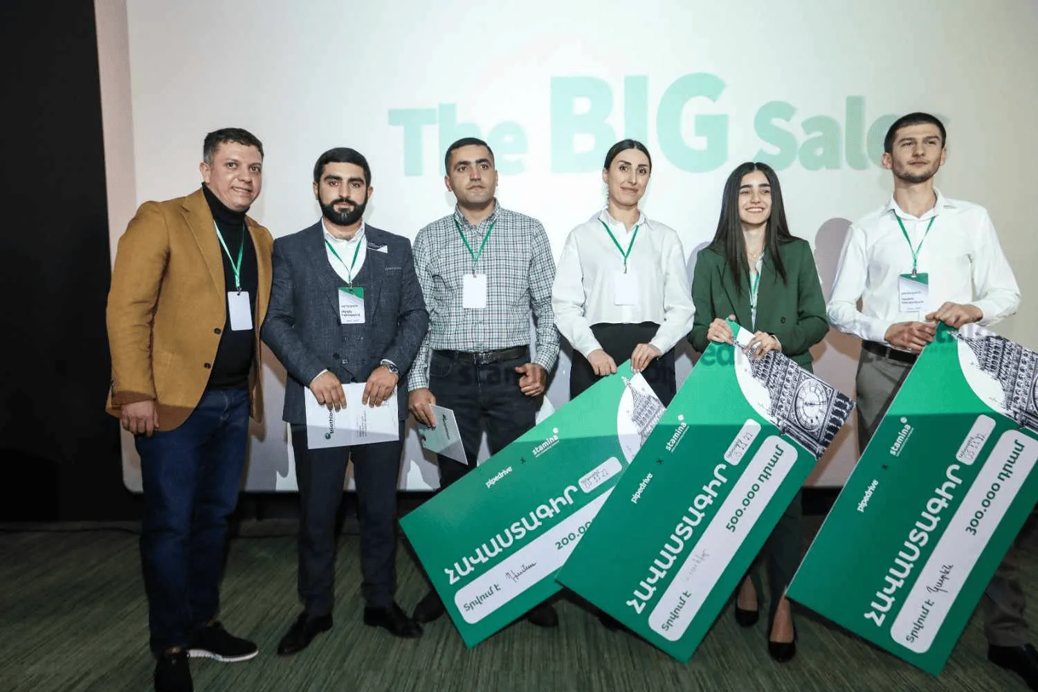 Five people standing side by side holding large green prize checks, with a projected sign behind them that reads 'The BIG sale.' Three of them hold checks with Armenian text and amounts of 200,000, 500,000, and 300,000 drams. They are wearing name badges and business casual attire.