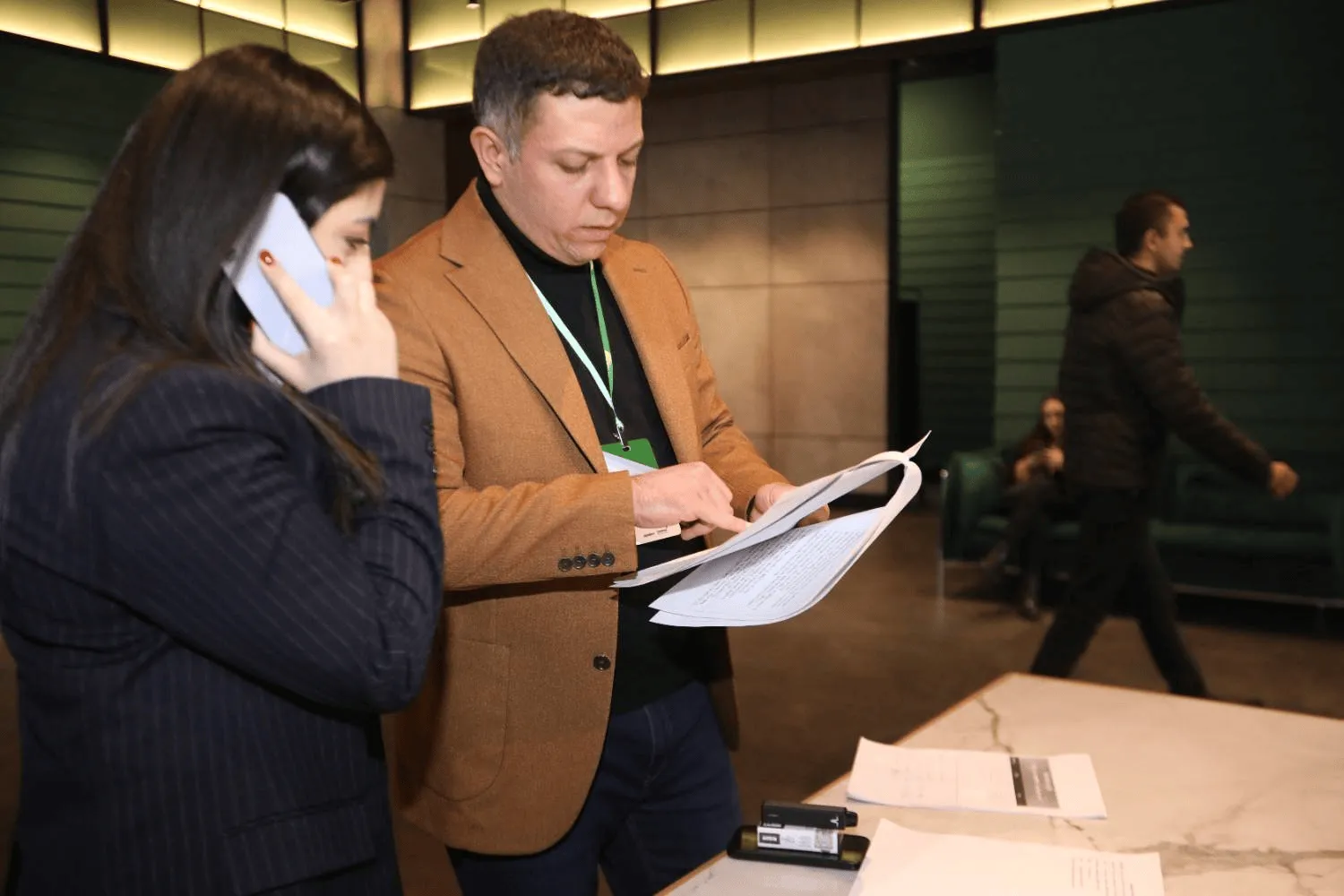 A man in a brown blazer and a woman in a dark striped jacket stand near a table with documents; the woman is talking on a phone while the man reviews papers. Two other people are visible in the background in a modern indoor setting.