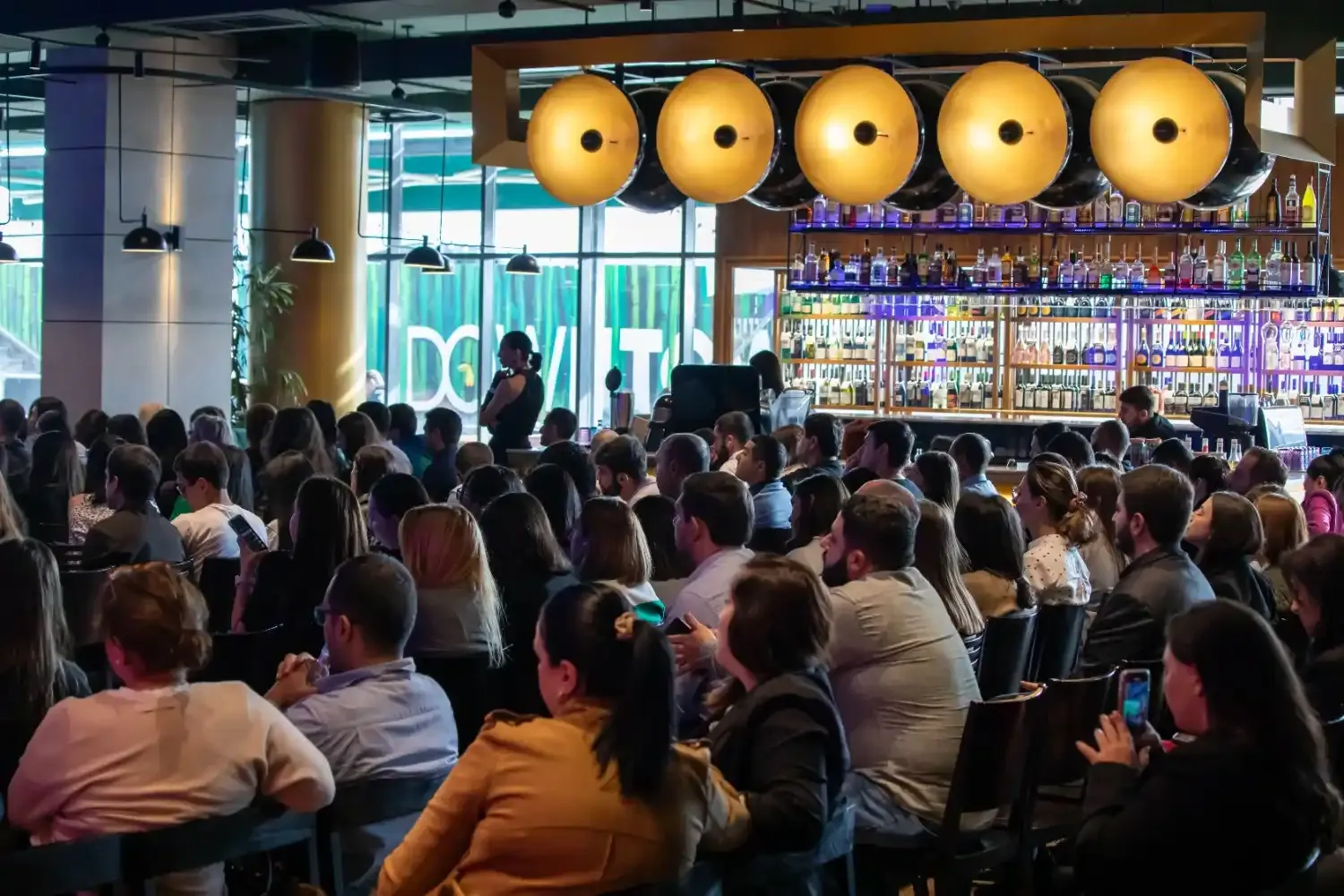 Large group of people seated in rows inside a modern venue with a bar in the background, focused on an event or speaker outside the frame.