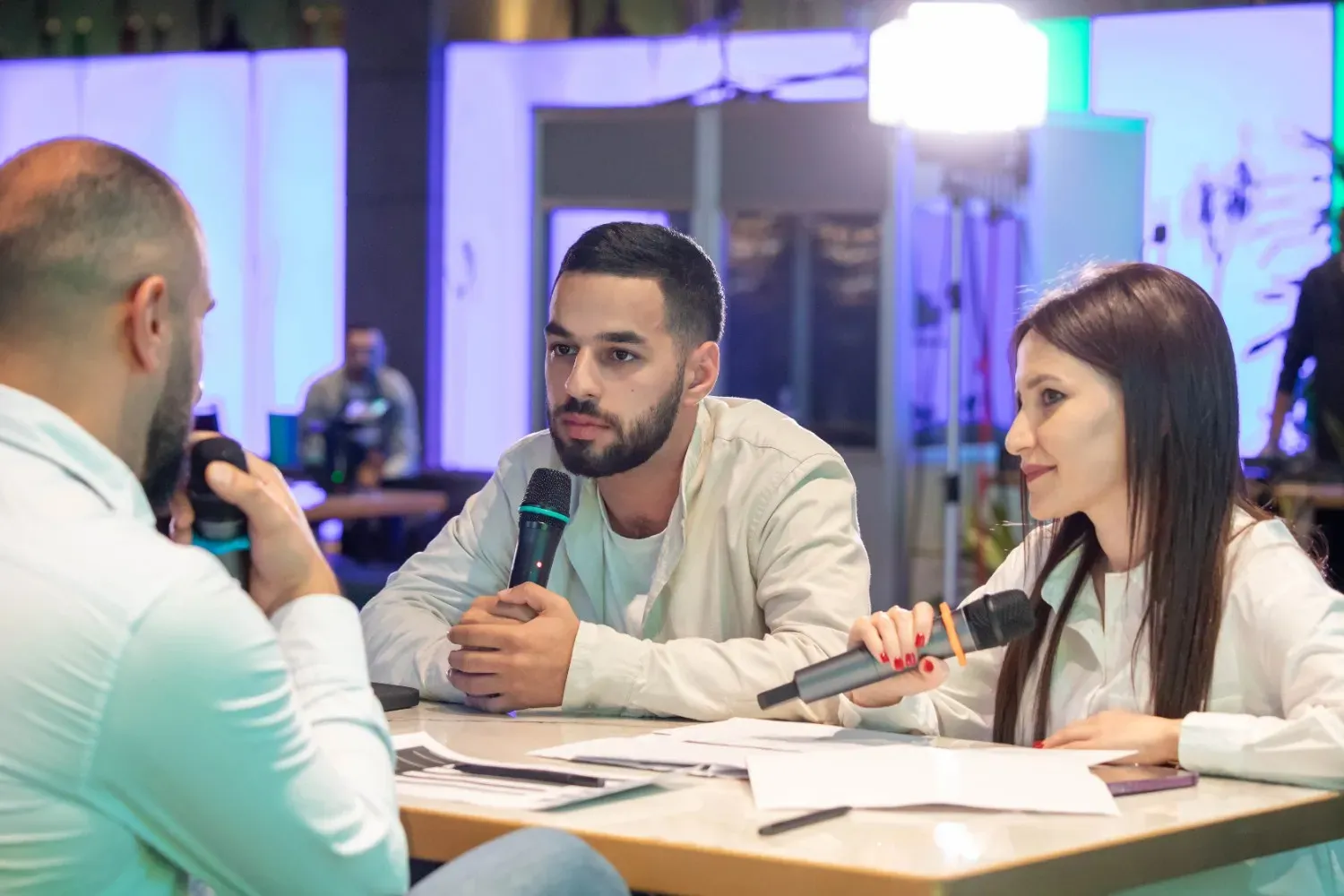 Three people seated around a table in a studio setting with blue and purple lighting; two men and one woman each holding microphones, engaged in a discussion, with papers and a smartphone on the table.