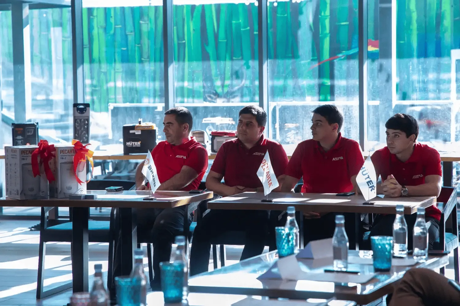 Four men in matching red polo shirts sit at a table in a bright room with large windows and bamboo-themed wallpaper, with product boxes tied with red and yellow ribbons and small branded flags on the table.
