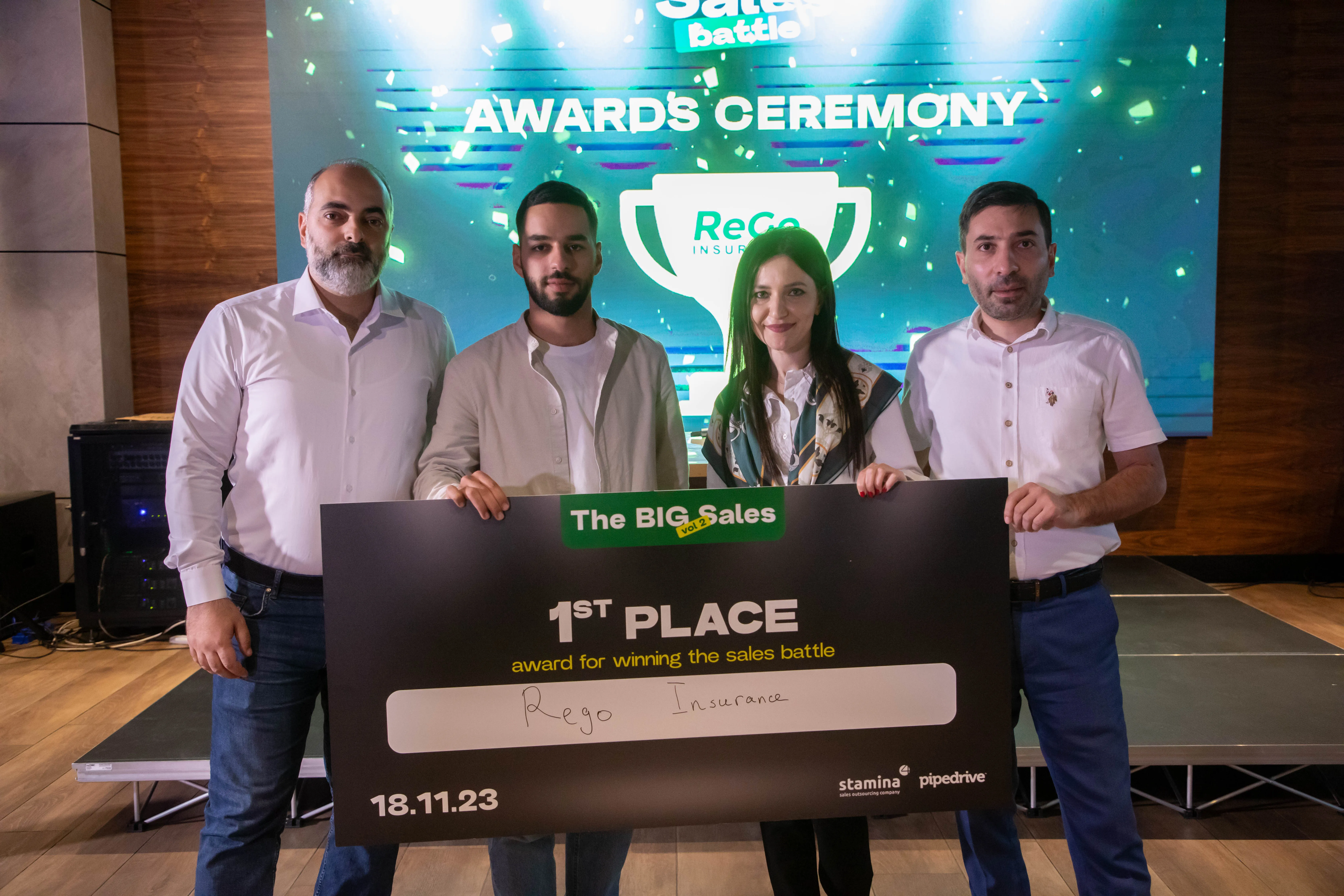 Four people standing indoors holding a large black award check that reads '1st Place award for winning the sales battle' with 'Rego Insurance' handwritten on it, dated 18.11.23, in front of a backdrop that says 'Awards Ceremony' with a trophy graphic.