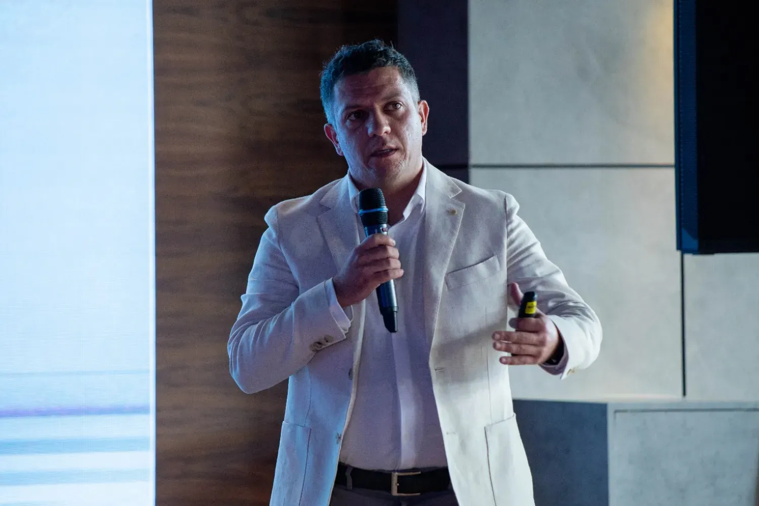 Man in a light-colored blazer and white shirt speaking into a microphone while holding a presentation remote, standing in front of a dark wood and light gray wall background.