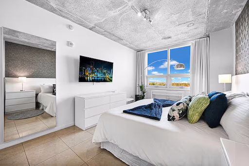 Modern bedroom with white walls, large window showing blue sky, bed with multiple pillows, wall-mounted TV, and a mirror reflecting another bed in an adjacent room.