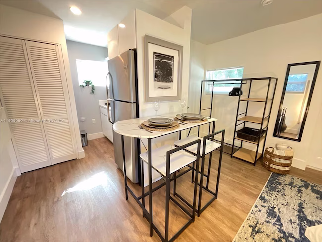 Modern small kitchen with wooden floor, stainless steel refrigerator, white marble top table set for two with black metal frame chairs, shelving unit, and narrow mirror on wall.
