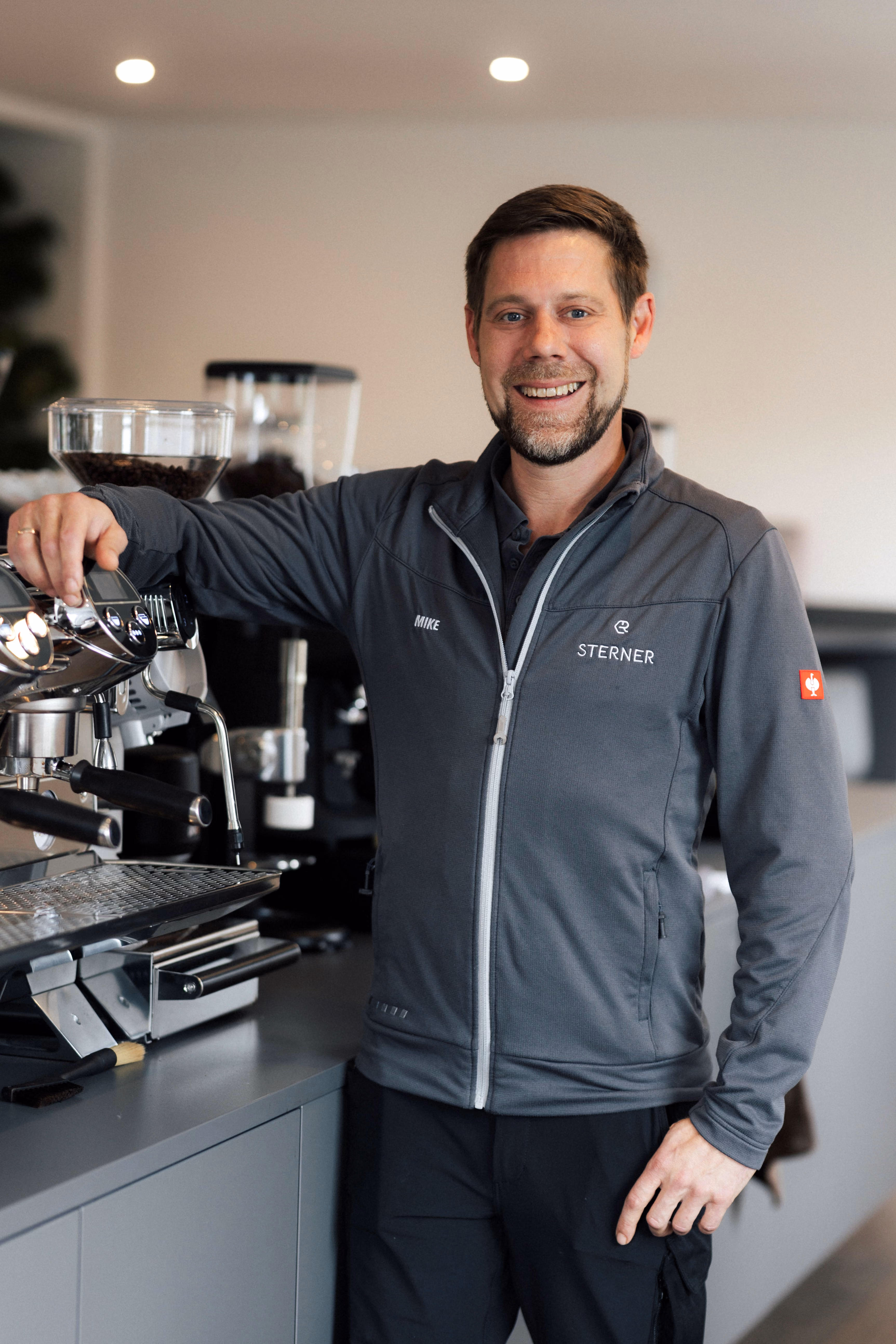 Lächelnder Mann lehnt an einer Espressomaschine in modernem Café, trägt graue Sterner-Jacke.