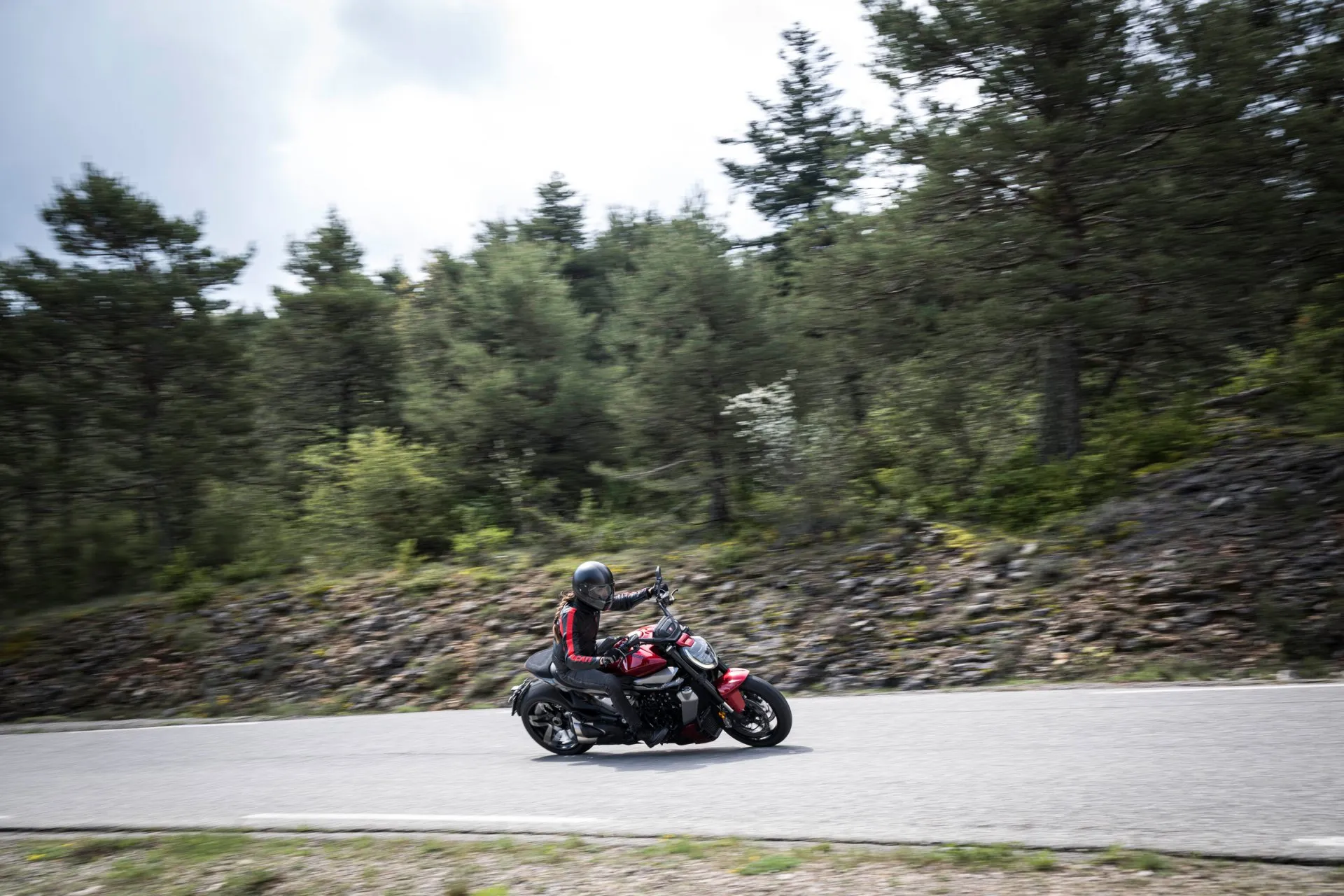 Motorradfahrer mit schwarzer Lederjacke fährt eine rote Ducati auf einer kurvigen Landstraße durch den Wald.