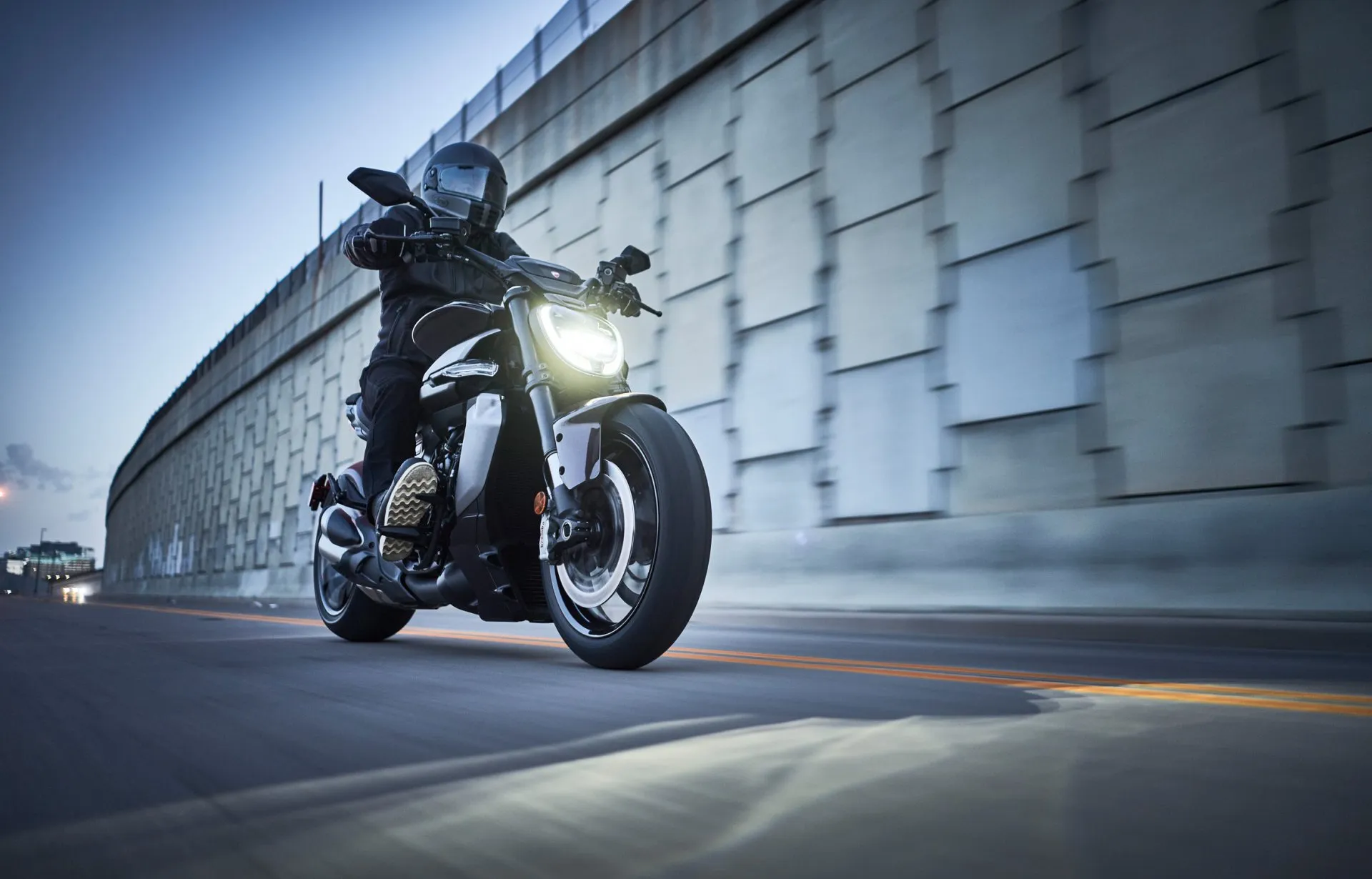 Motorradfahrer auf einer schwarzen Ducati fährt bei Dämmerung entlang einer Mauer in der Stadt.