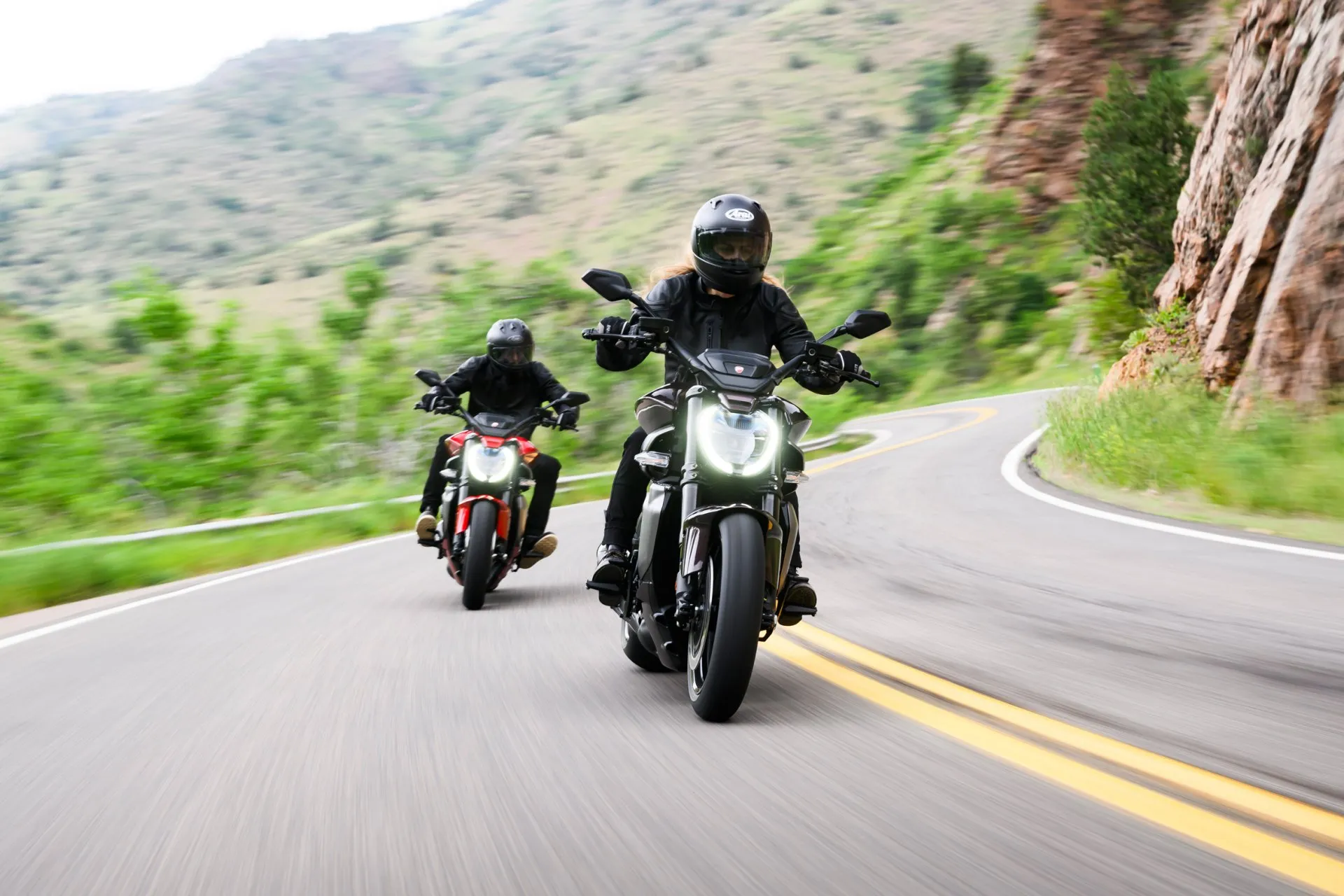 Zwei Motorradfahrer auf Ducati-Maschinen fahren eine kurvige Bergstraße entlang.