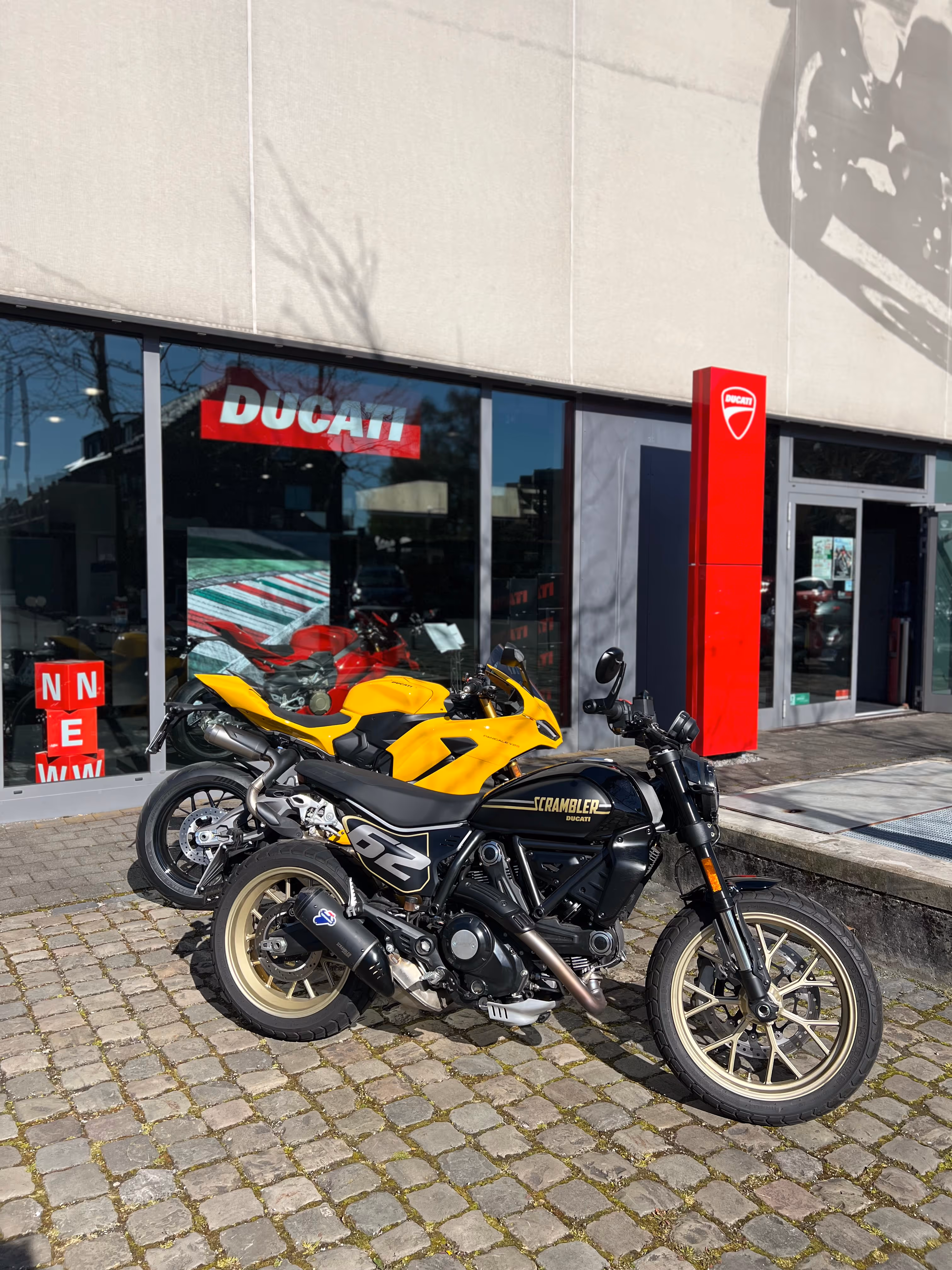 Zwei Ducati-Motorräder, ein gelbes Sportmodell und ein schwarzes Scrambler-Modell, vor einem Ducati-Händler mit roten Logos.