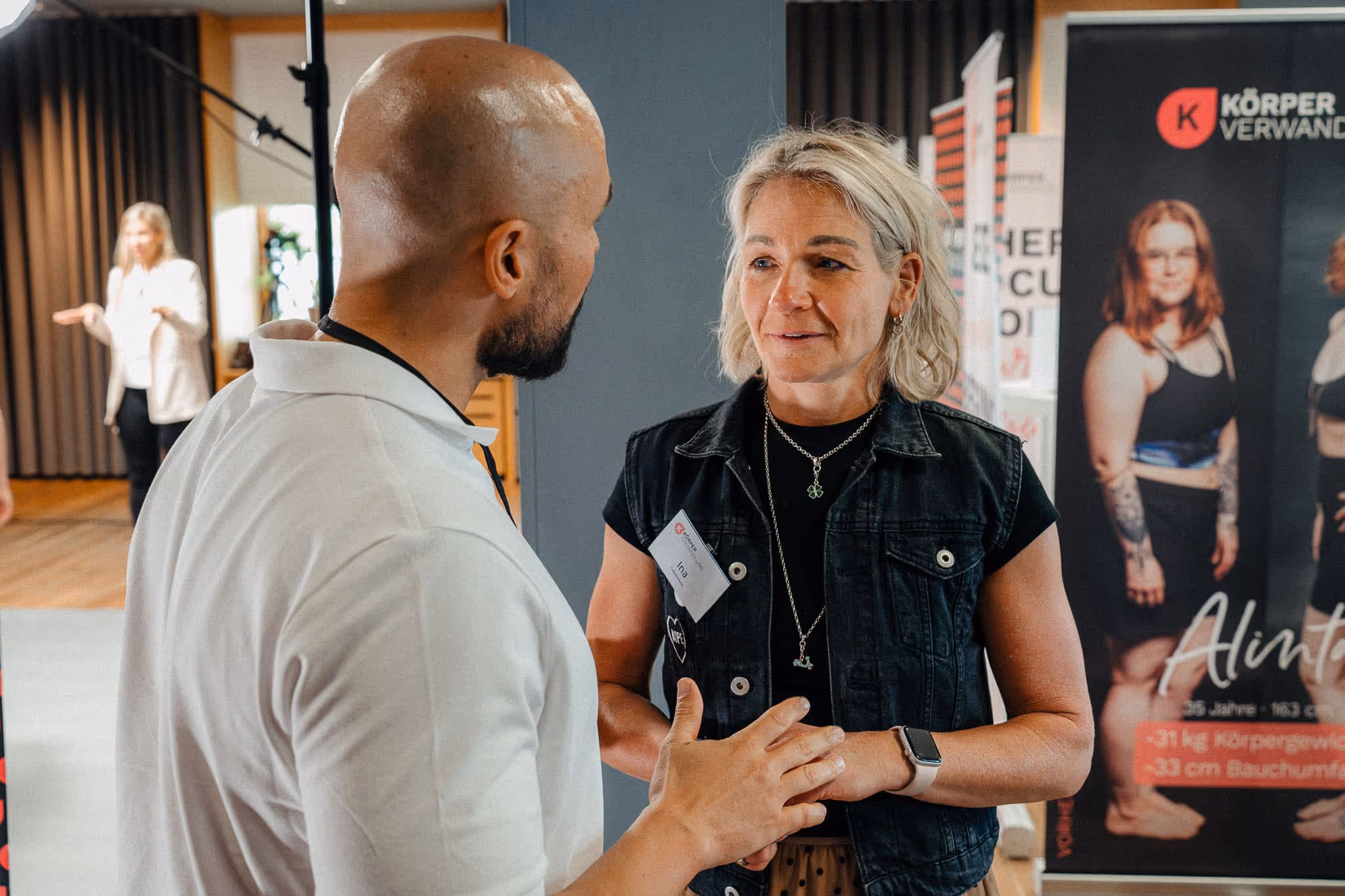 Ernährungsexperte Andreas Kotte im Gespräch mit einer Frau während einer persönlichen Ernährungsberatung bei Körperverwandlung. Im Hintergrund sind Erfolgsgeschichten von Teilnehmerinnen zu sehen.
