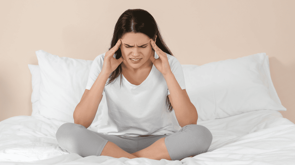 Frau sitzt erschöpft auf dem Bett und hält ihren Kopf – typische körperliche und emotionale Belastung durch hormonelle Verhütung.