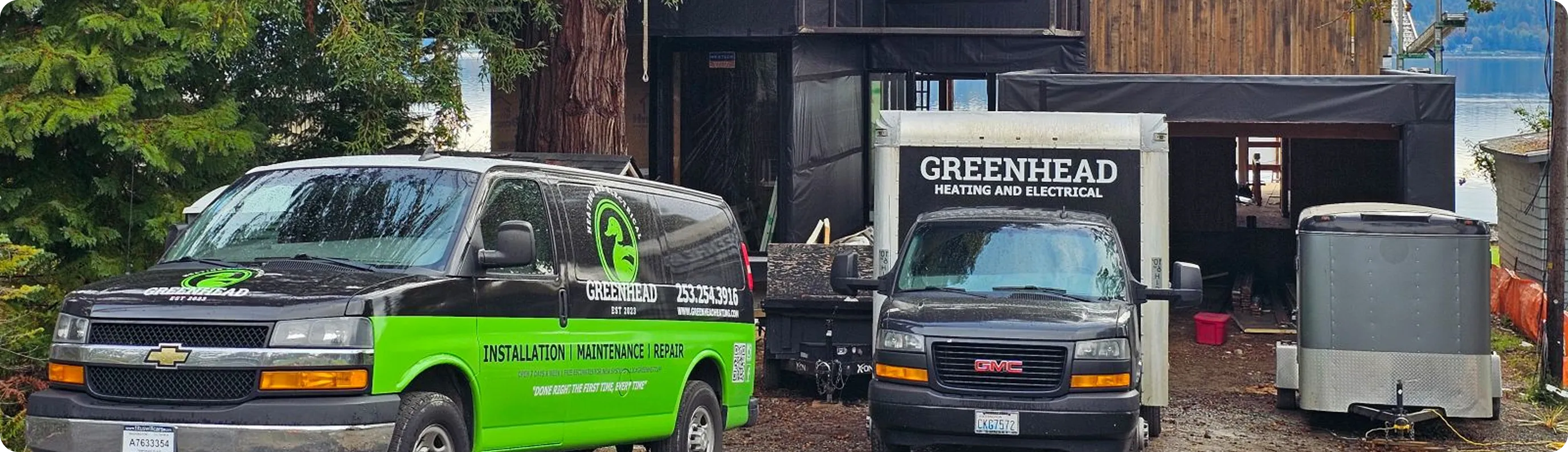 Greenhead Heating and Electrical trucks parked outside a residential construction site surrounded by trees.
