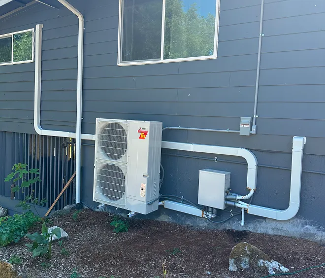 Exterior view of an air conditioning unit installed on a home's wall, with visible piping and landscaping.
