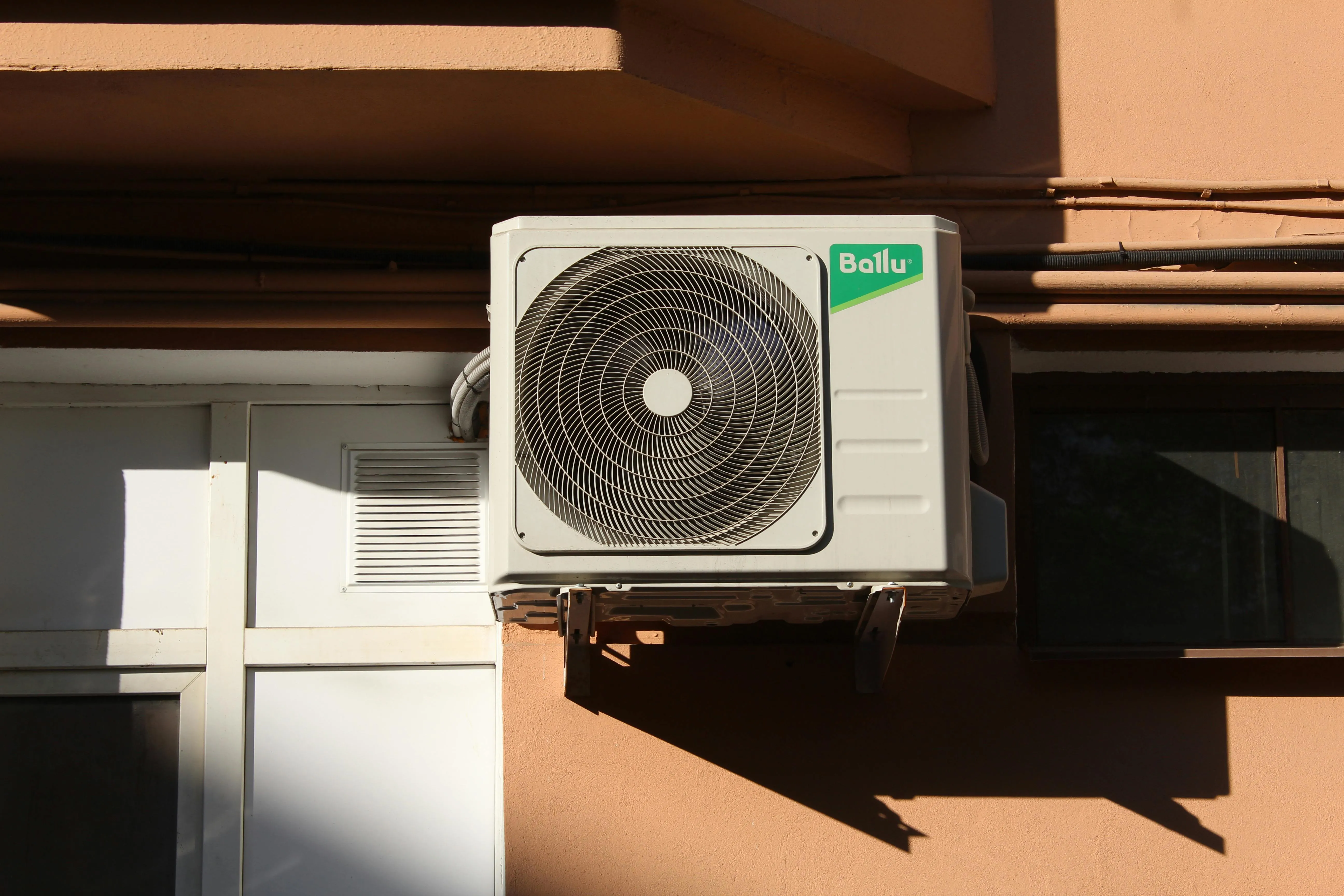 Wall-mounted Ballu air conditioner unit installed next to a window on a building exterior.
