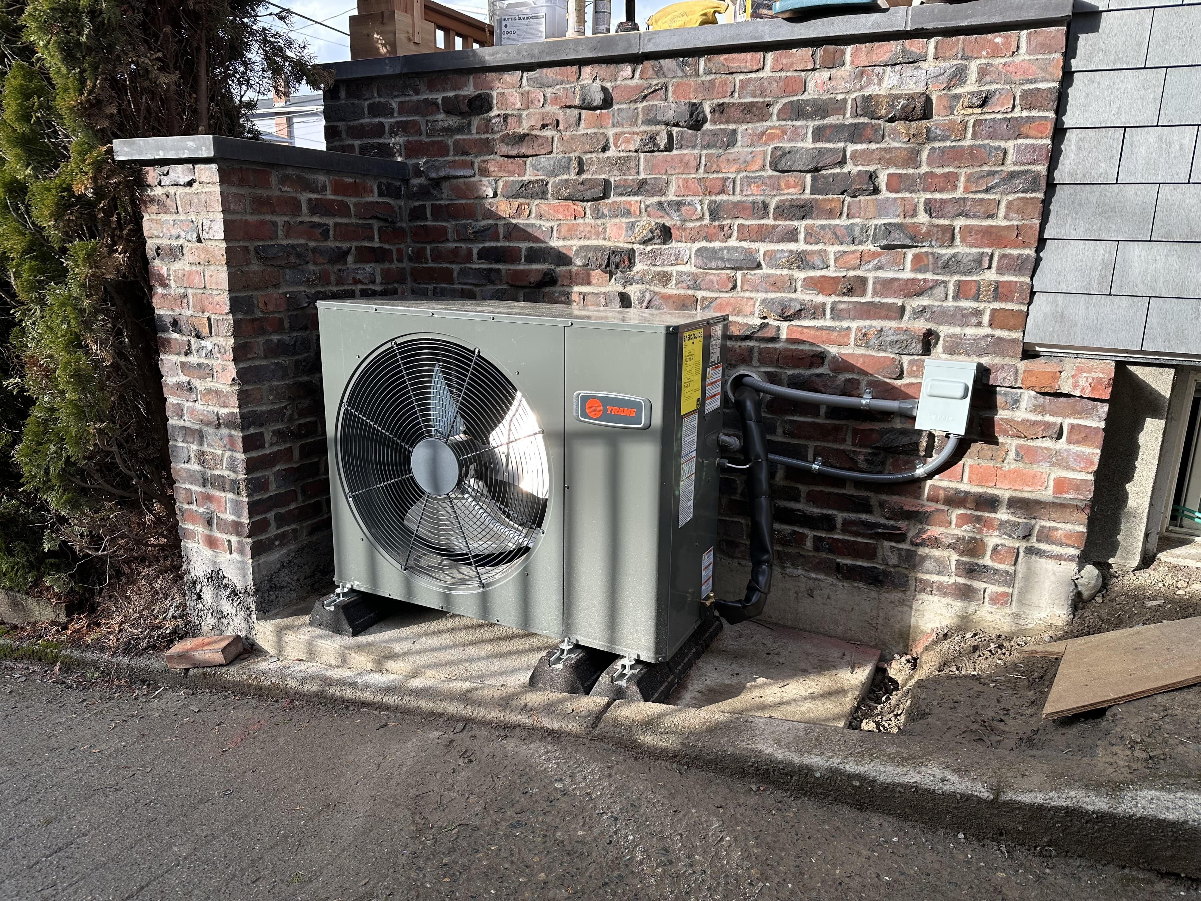 A Trane air conditioning unit installed next to a brick wall in a residential setting.
