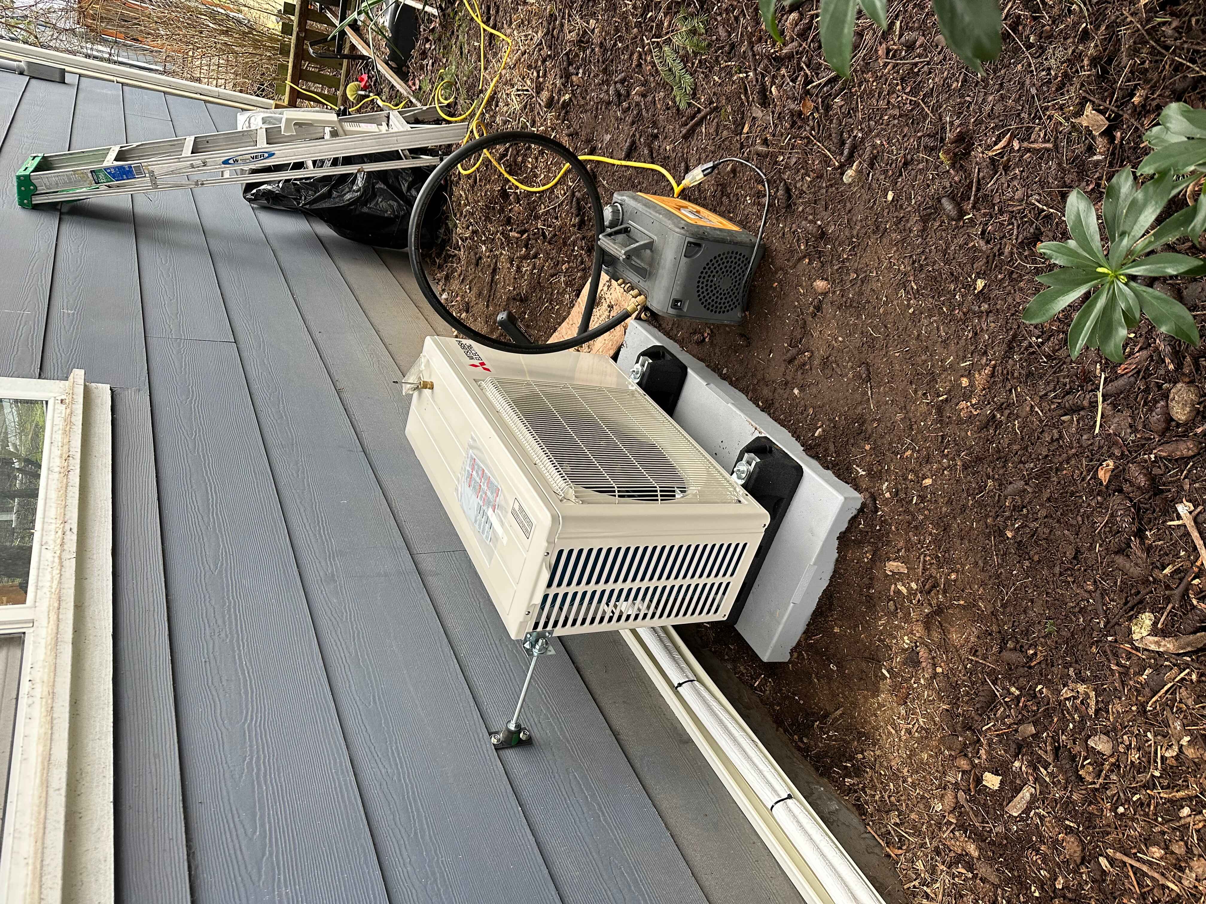 Outdoor air conditioning unit installation next to the house, with a ladder and surrounding landscaping.
