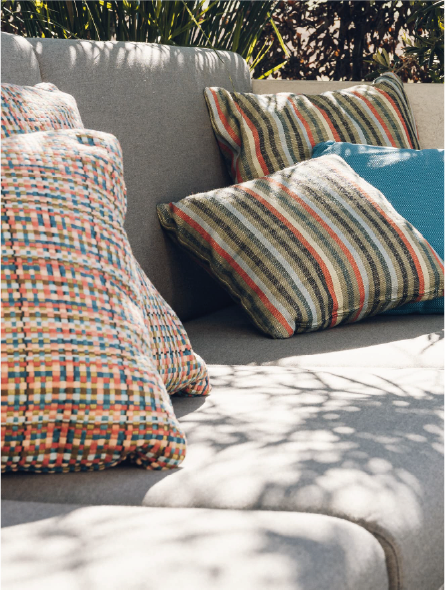 Gray outdoor sofa with colorful striped and patterned throw pillows under dappled sunlight.