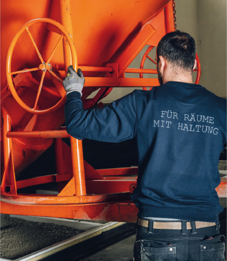 Worker in a navy sweatshirt operating an orange industrial machine with large handwheels.