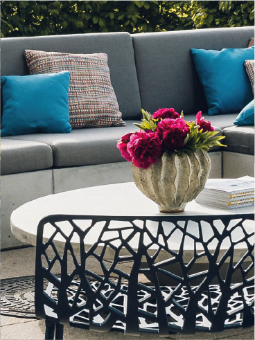 Outdoor seating with gray couch, colorful pillows, white table with vase of pink flowers, and black patterned chair.
