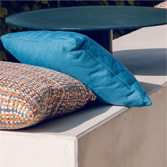Two outdoor cushions, one blue and one patterned, placed on a white concrete bench under a round green table.