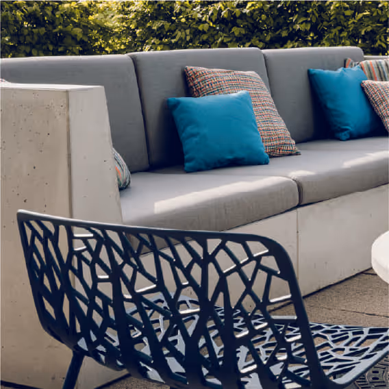 Outdoor concrete sectional sofa with gray cushions and blue and patterned pillows beside a black lattice chair.