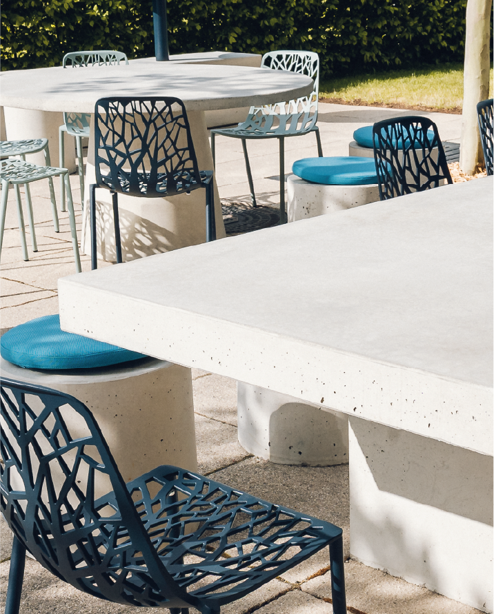 Outdoor concrete tables with artistic metal chairs and round stools topped with blue cushions.