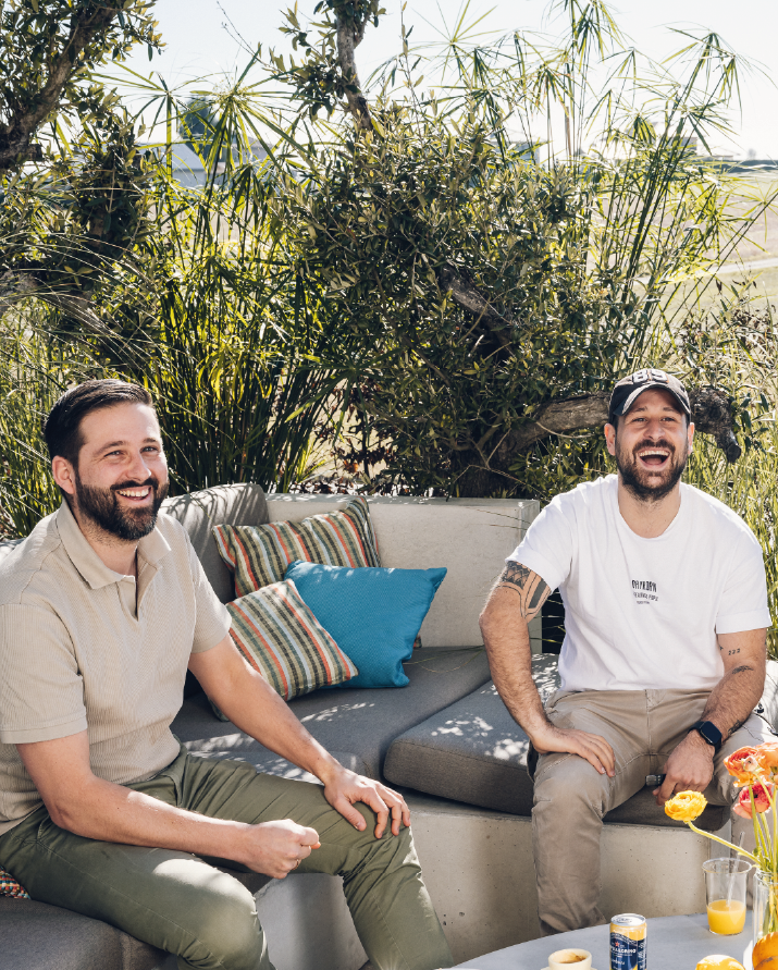 Two men laughing and sitting on a patio couch with colorful pillows under green plants.