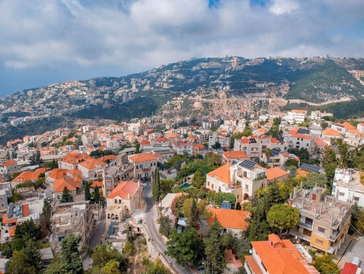 Lebanon town with dense houses having terracotta roofs surrounded by greenery under a partly cloudy sky.