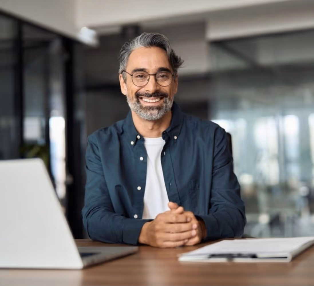 Smiling gray-haired man with glasses sitting at desk with laptop and notebook in office.