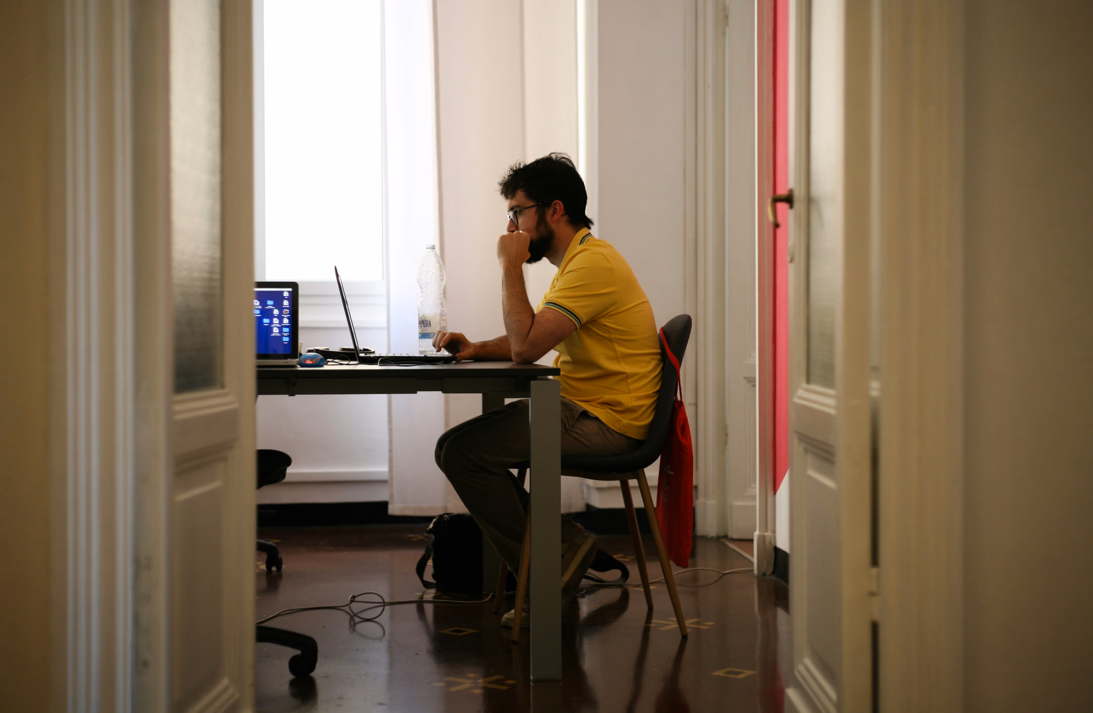 Man in yellow shirt sitting at a desk working on a laptop in a room with a window behind him.