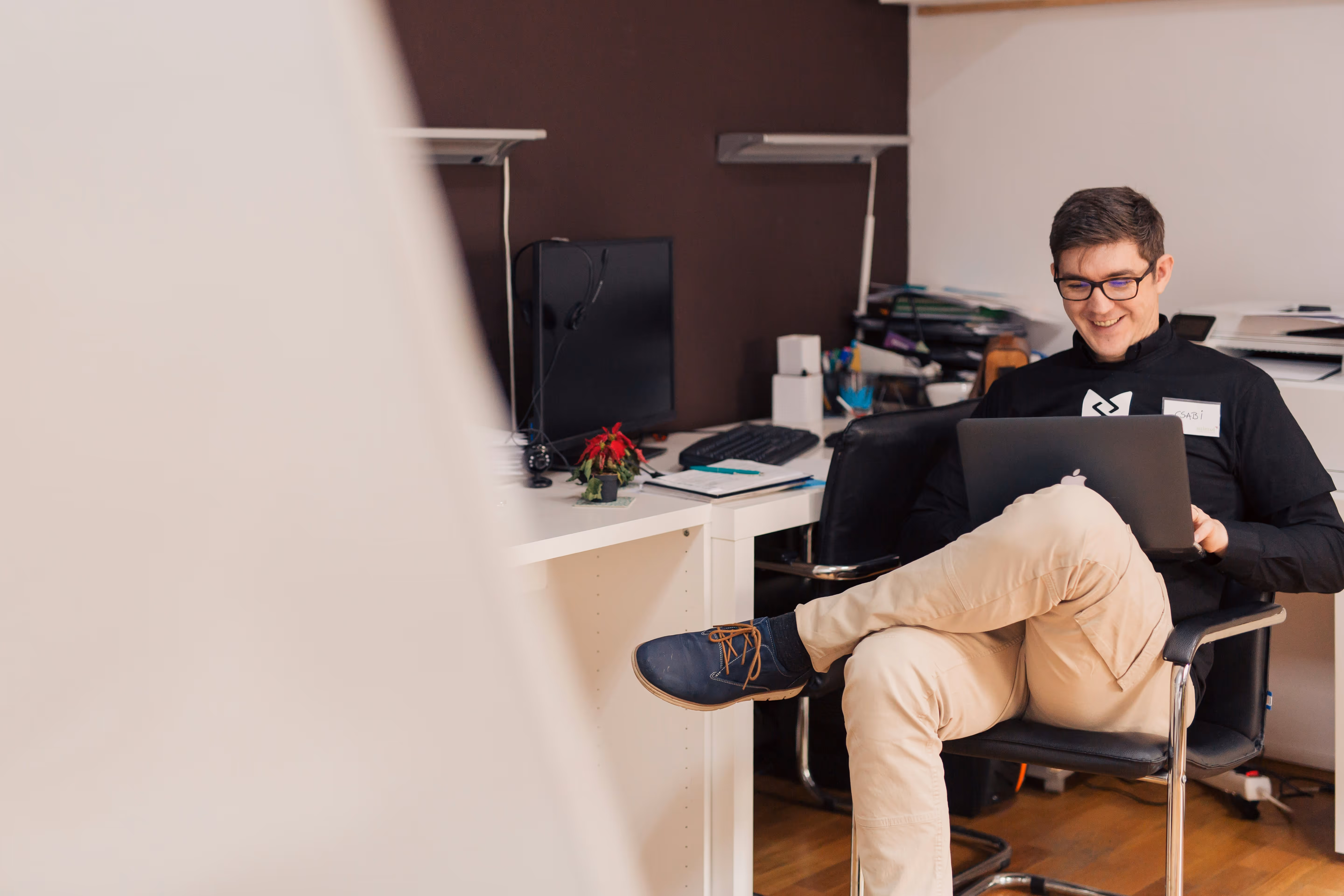 Young man with glasses smiling while sitting cross-legged on an office chair using a laptop.