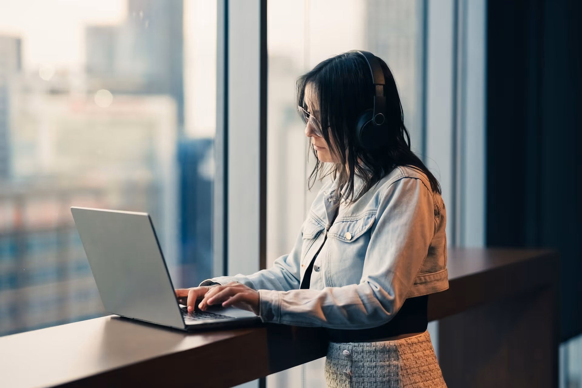Woman wearing headphones and glasses typing on a laptop by a large window in an office.