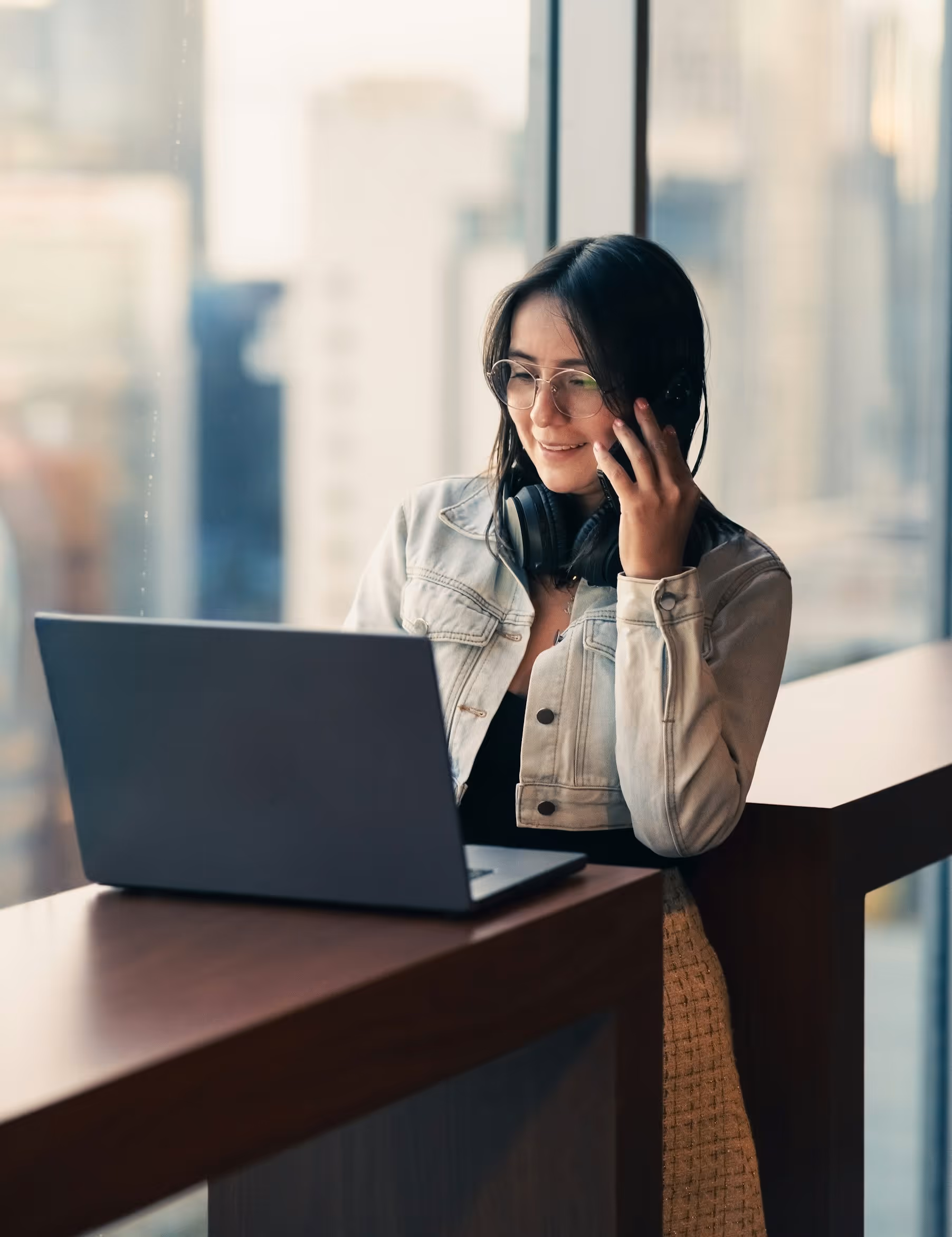 Woman in glasses and denim jacket using laptop and talking on phone by large window.