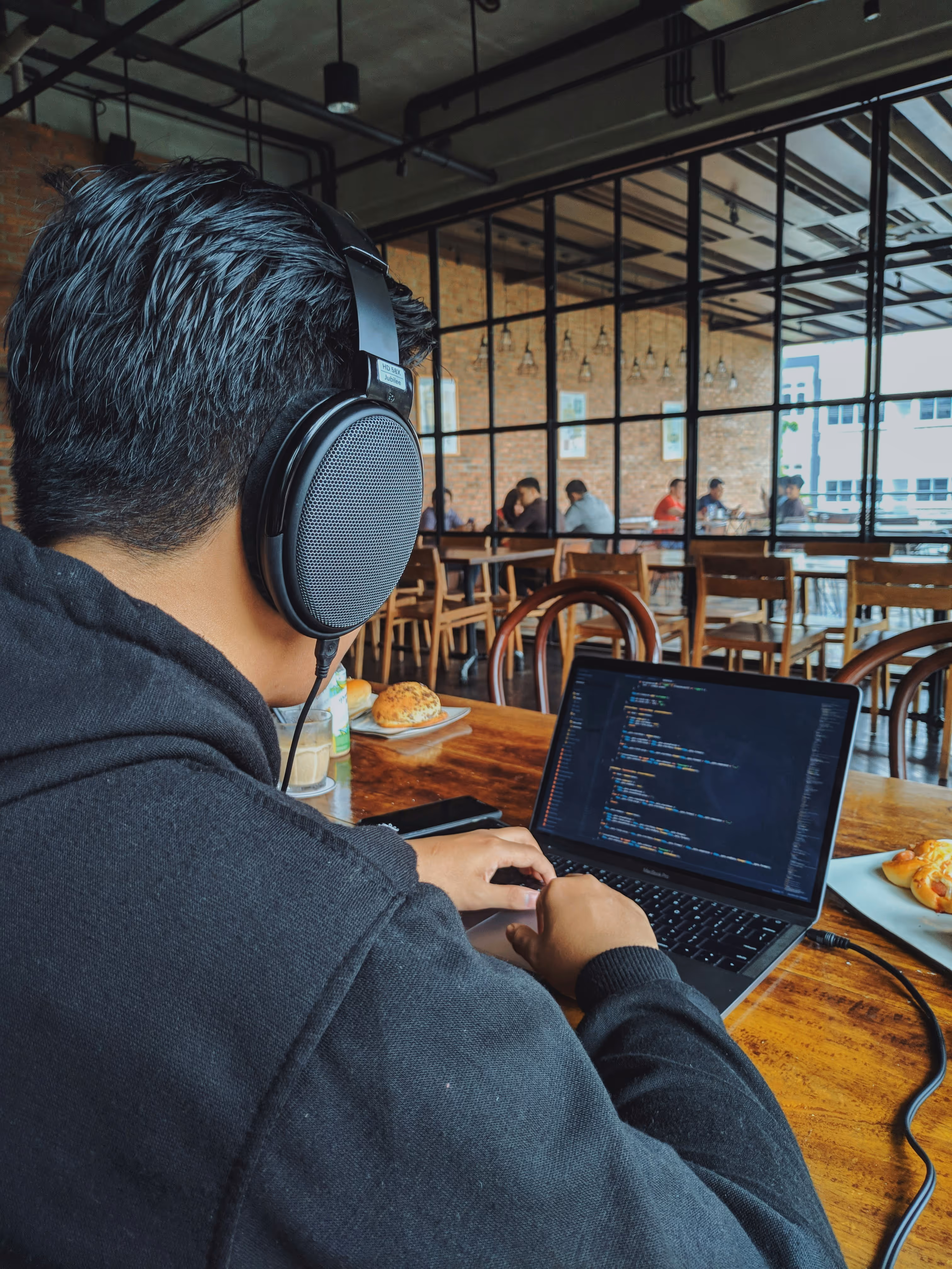 Person wearing large headphones working on a laptop with code displayed in a cafe.