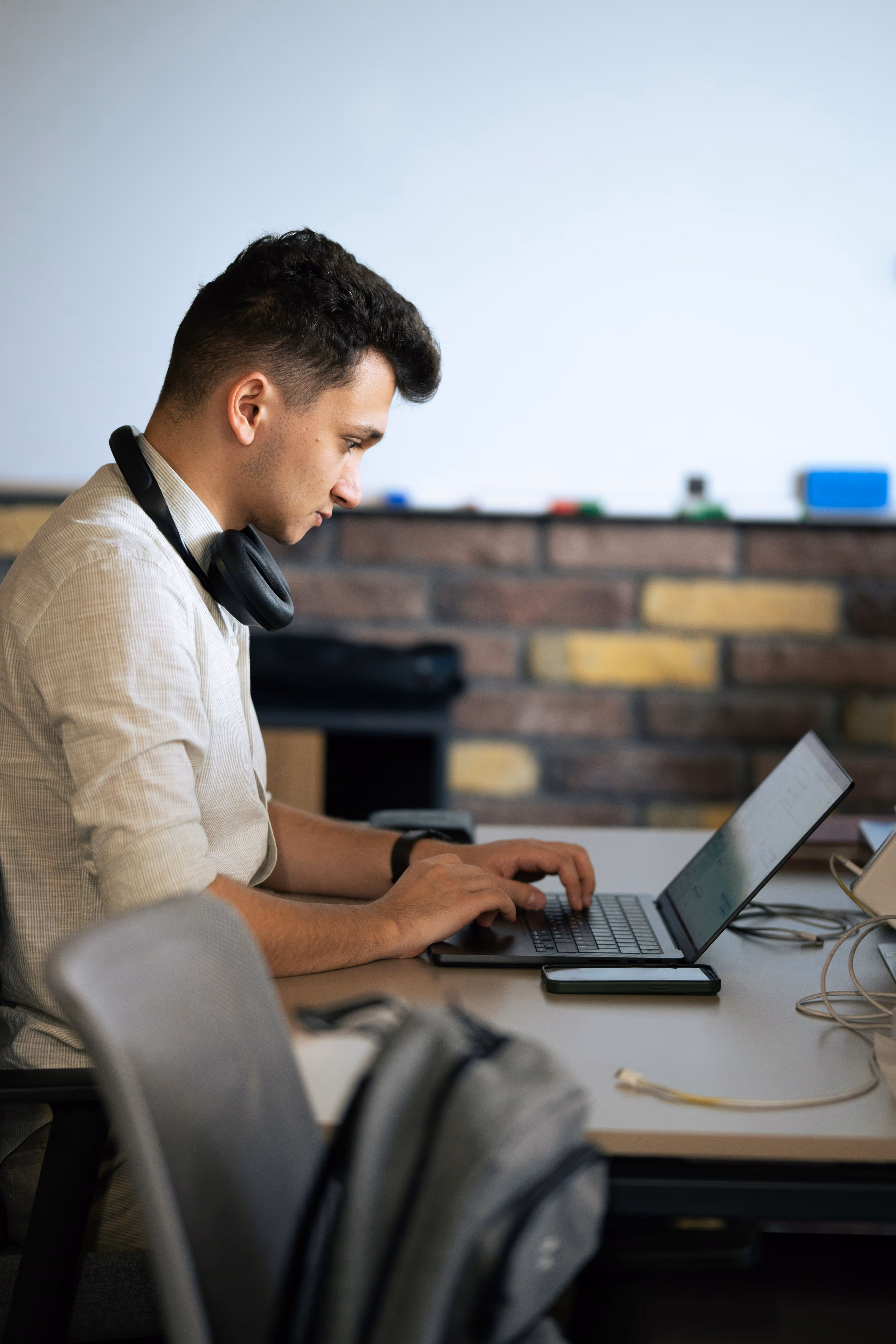Young man with headphones typing on a laptop at a desk in an office setting.