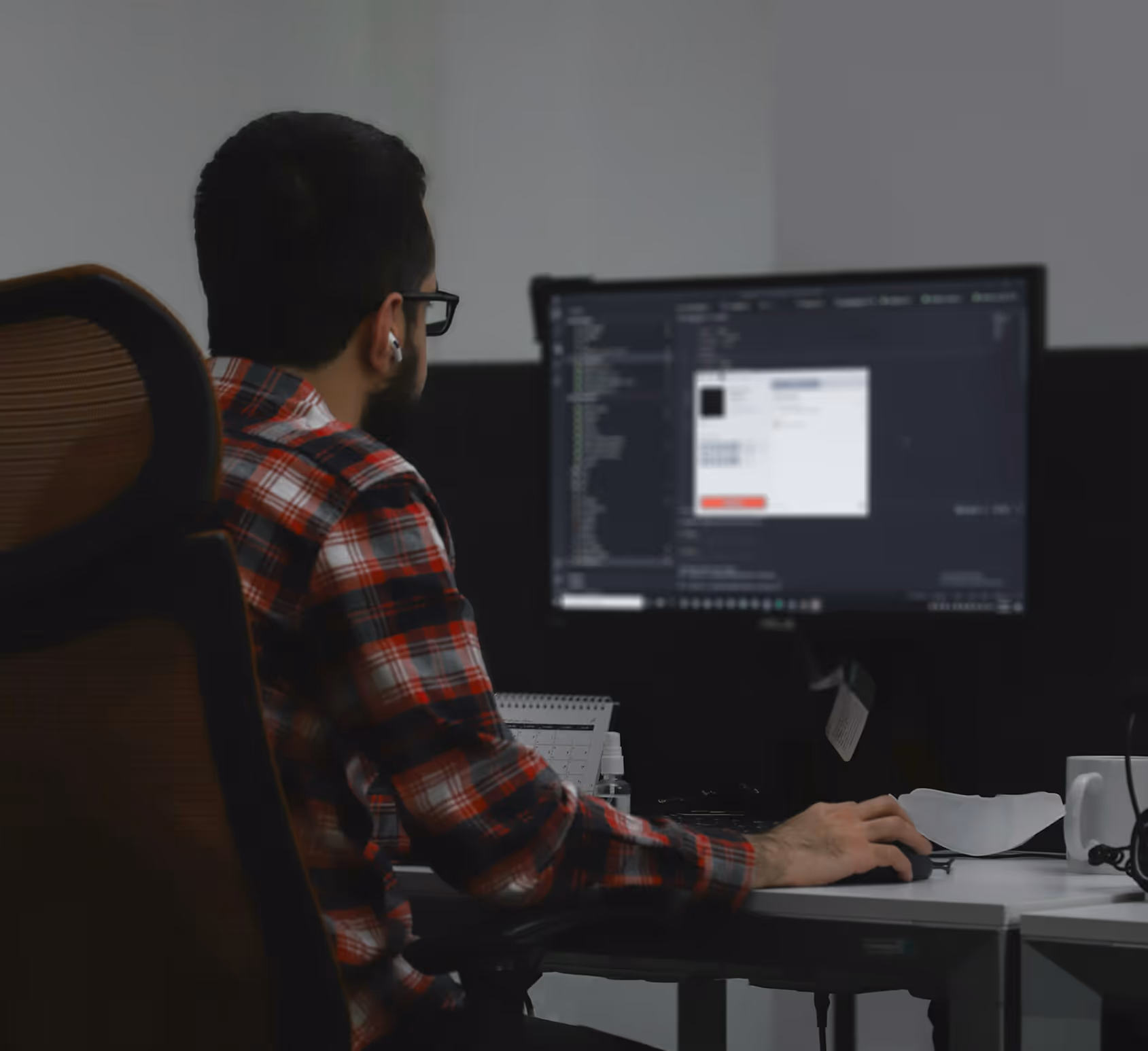 Man wearing glasses and red plaid shirt working on a computer with code displayed on the monitor.