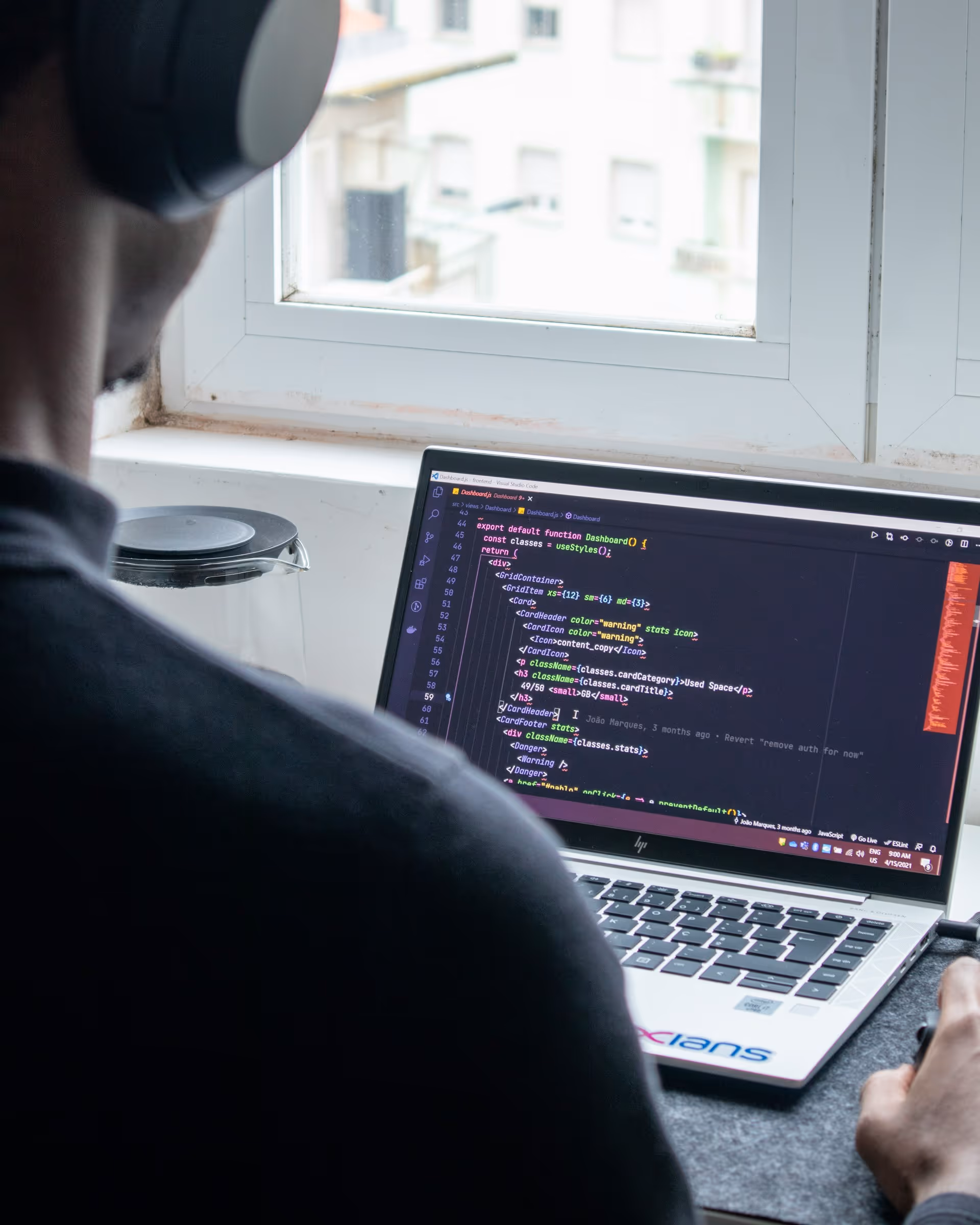 Person wearing headphones coding on a laptop displaying JavaScript code near a window.