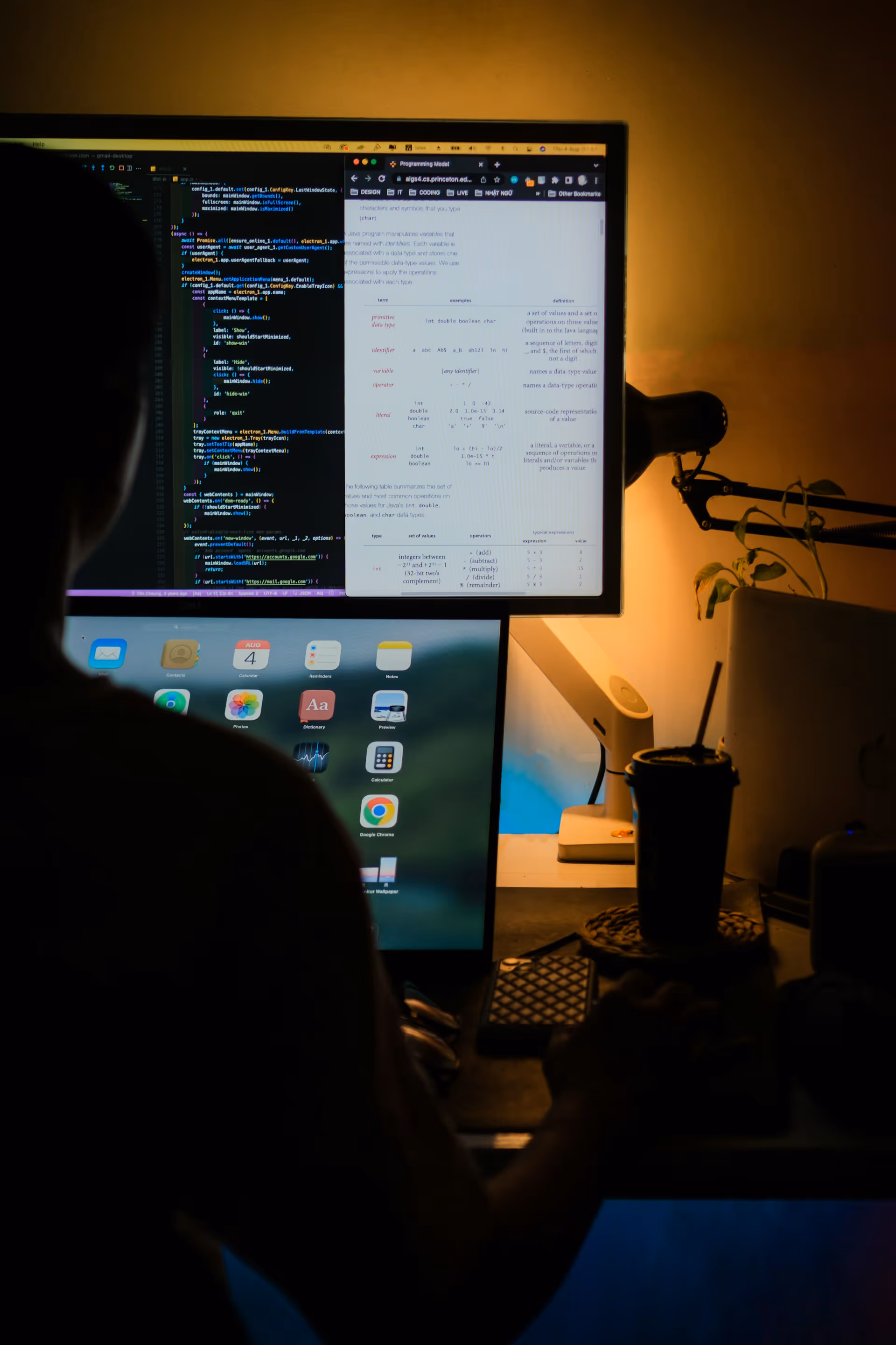 Person working at a desk with two computer screens showing coding and app icons in a dimly lit room.