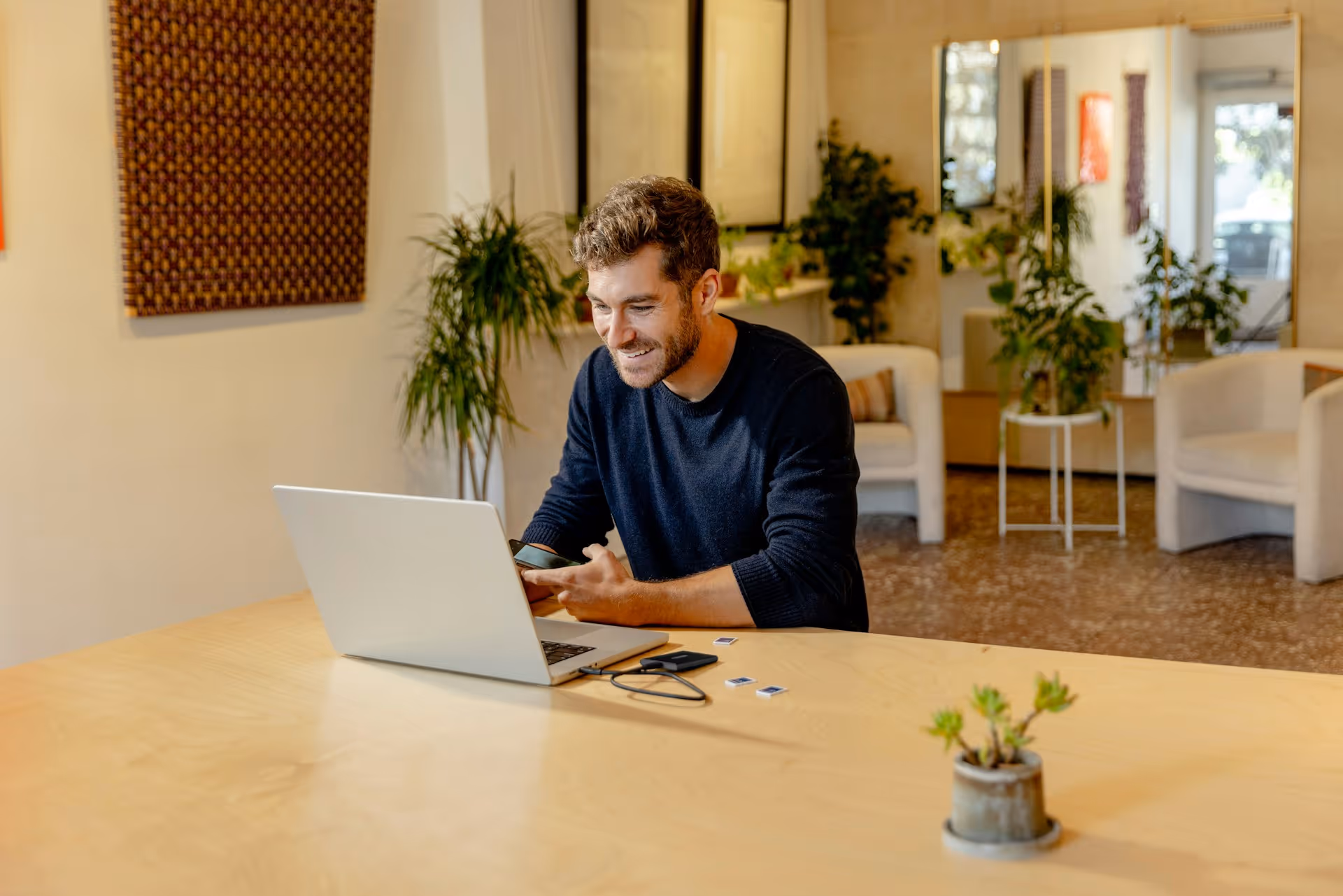 Man in black sweater smiling working on laptop at a wooden desk in a modern room with plants.