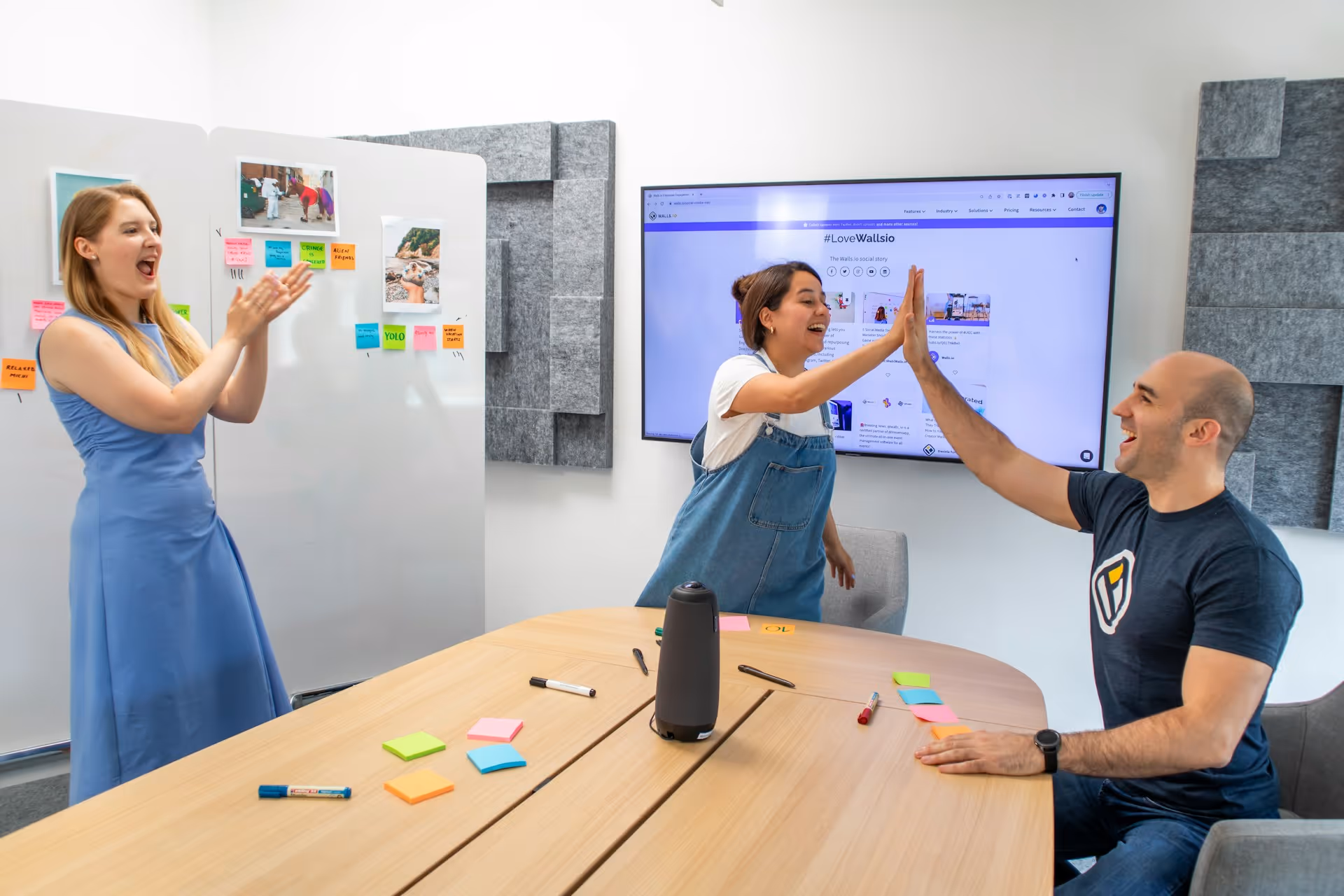 Two colleagues in a meeting room giving each other a high five while a third person claps and smiles.