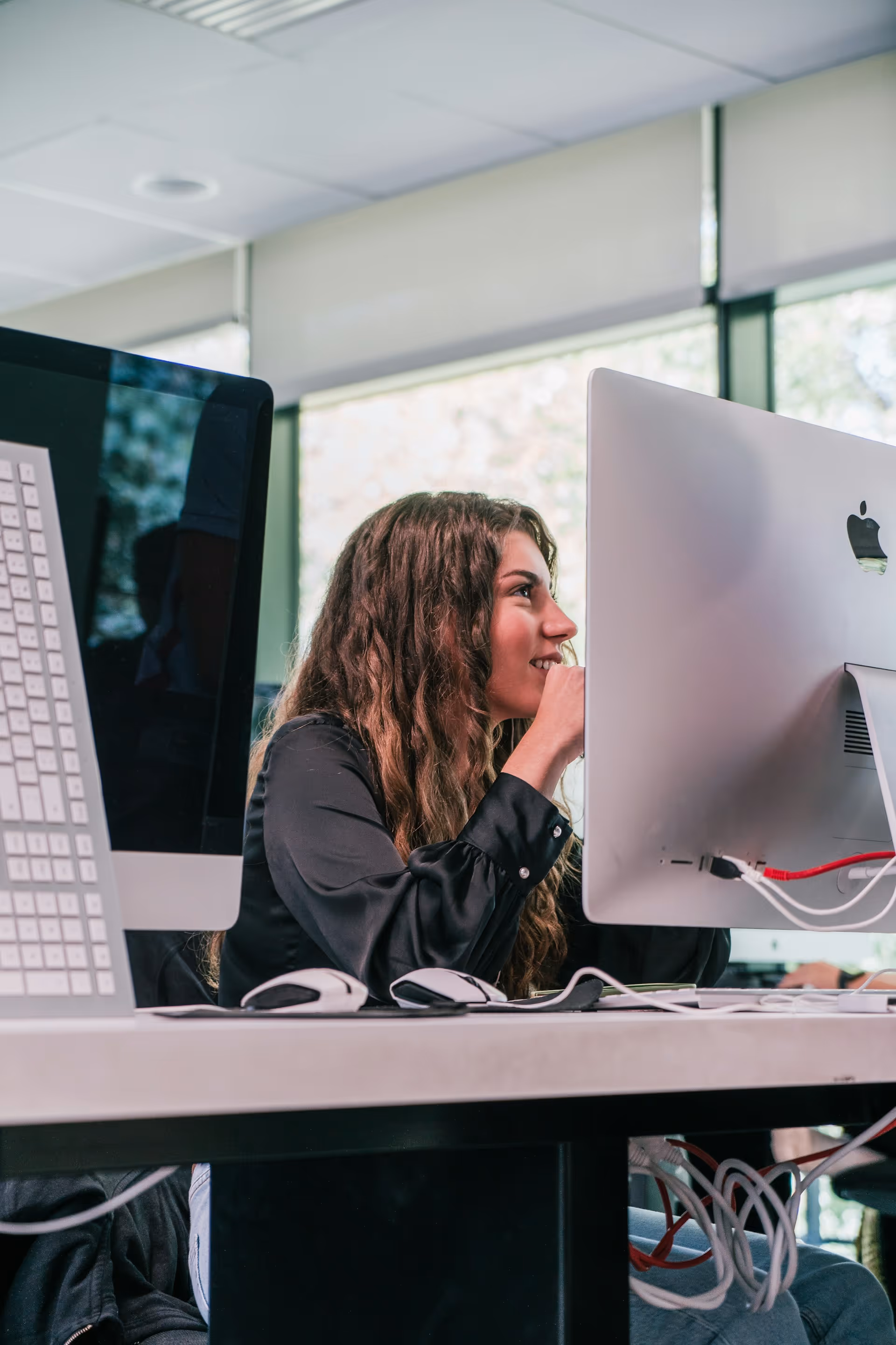 A woman with long curly hair sitting at a desk working on an Apple desktop computer in an office.