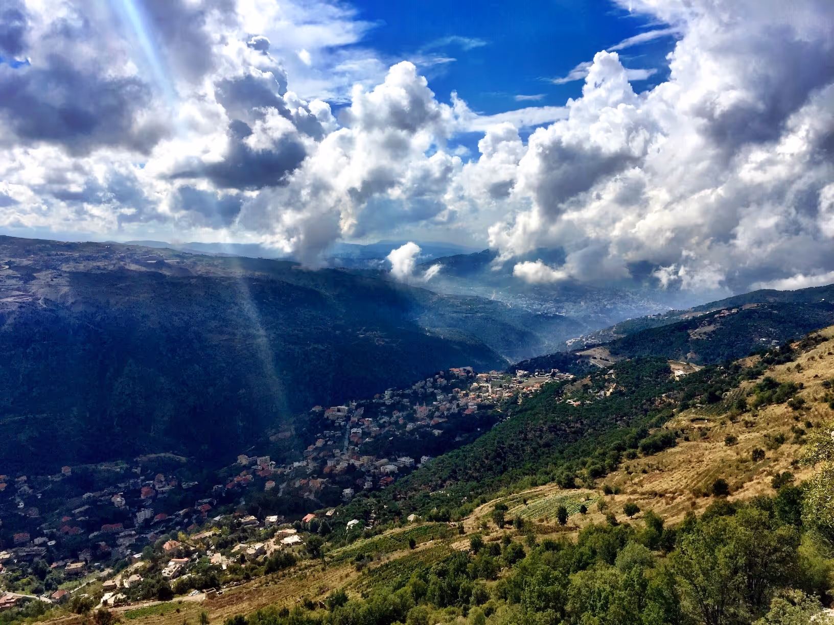 Mountain valley with scattered houses under a blue sky filled with large white clouds and sun rays.