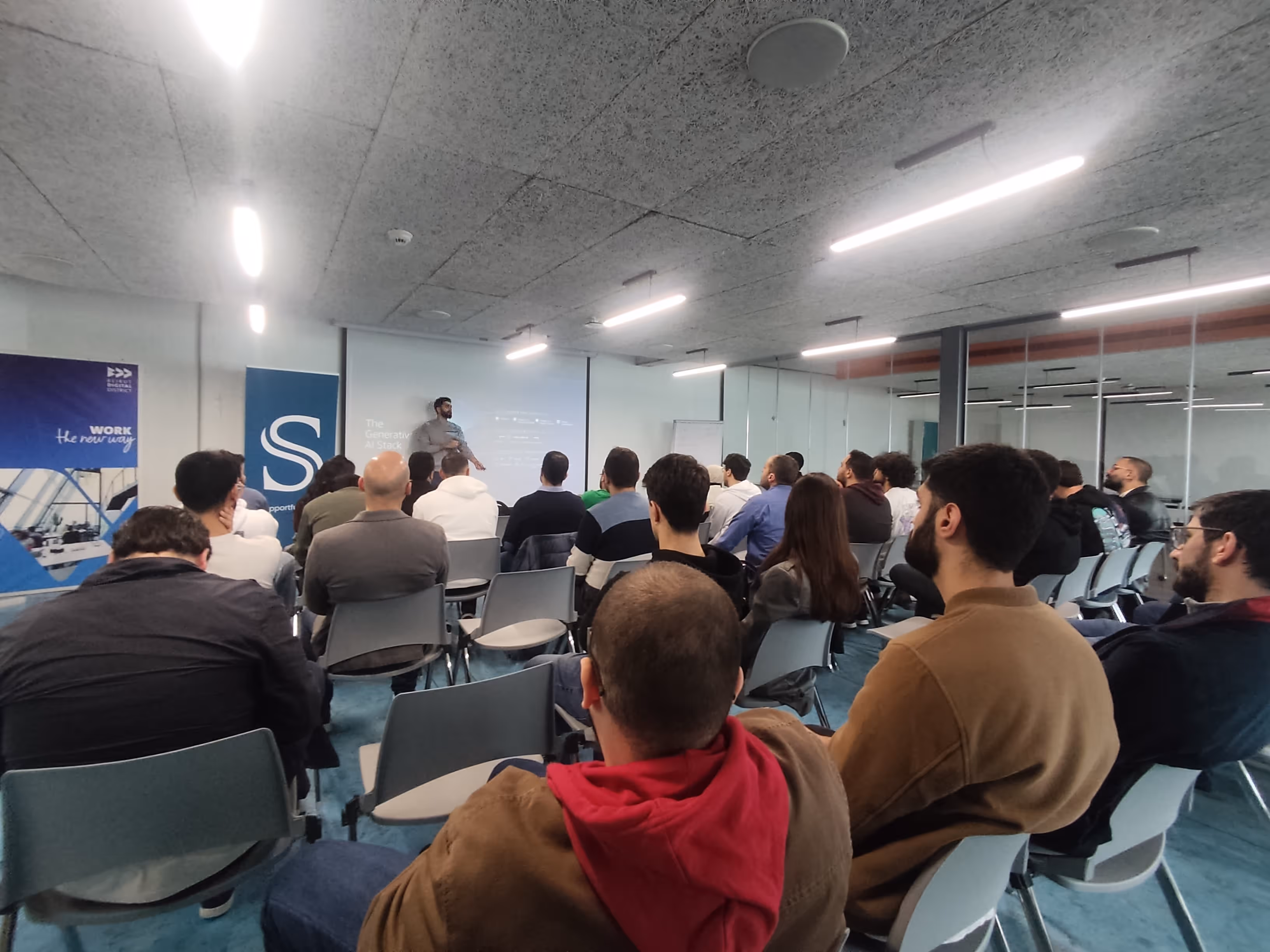 Audience seated facing a presenter explaining 'The Generative AI Stack' projected on a screen in a modern conference room.