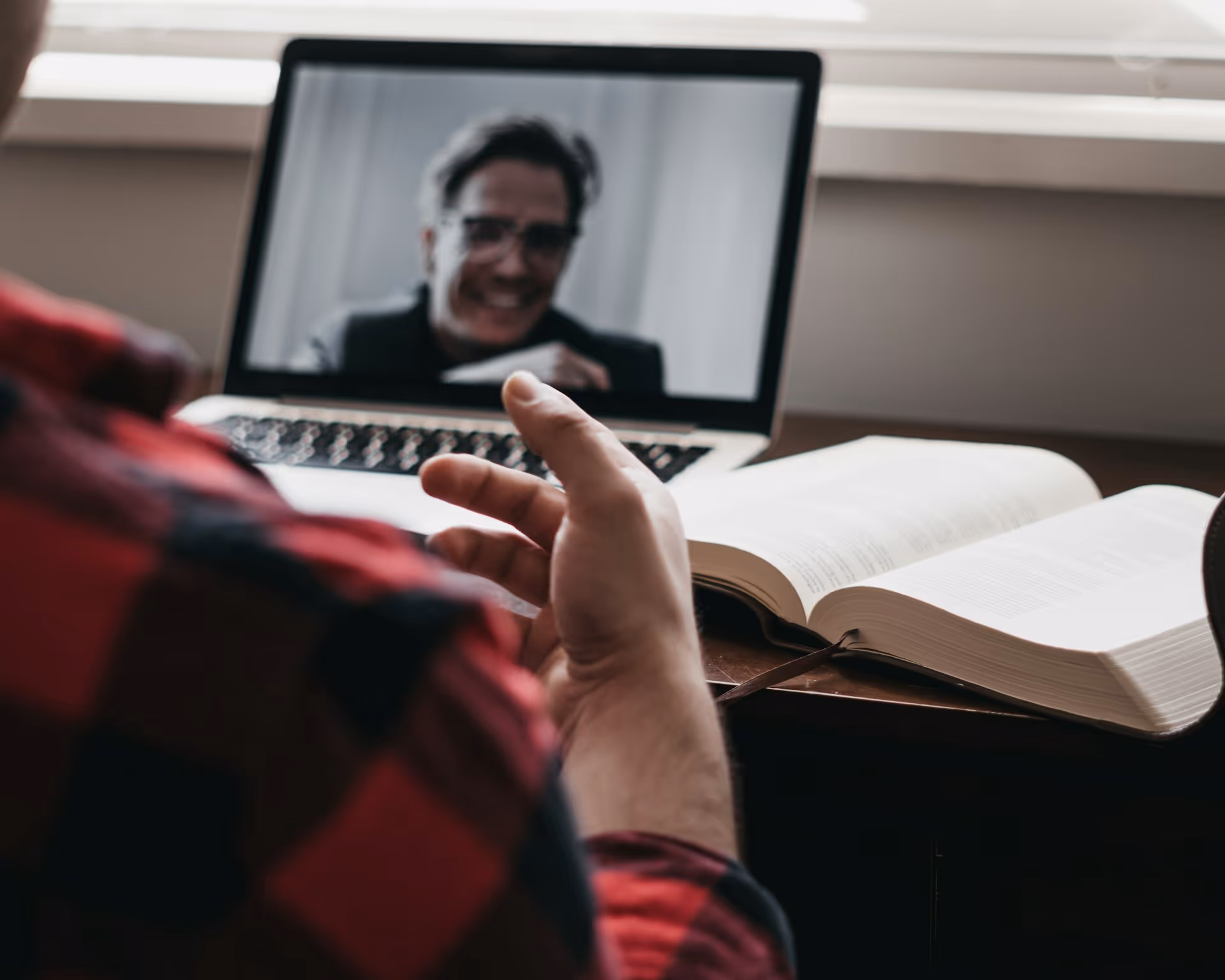 Person in red plaid shirt video conferencing on laptop with open book on desk.