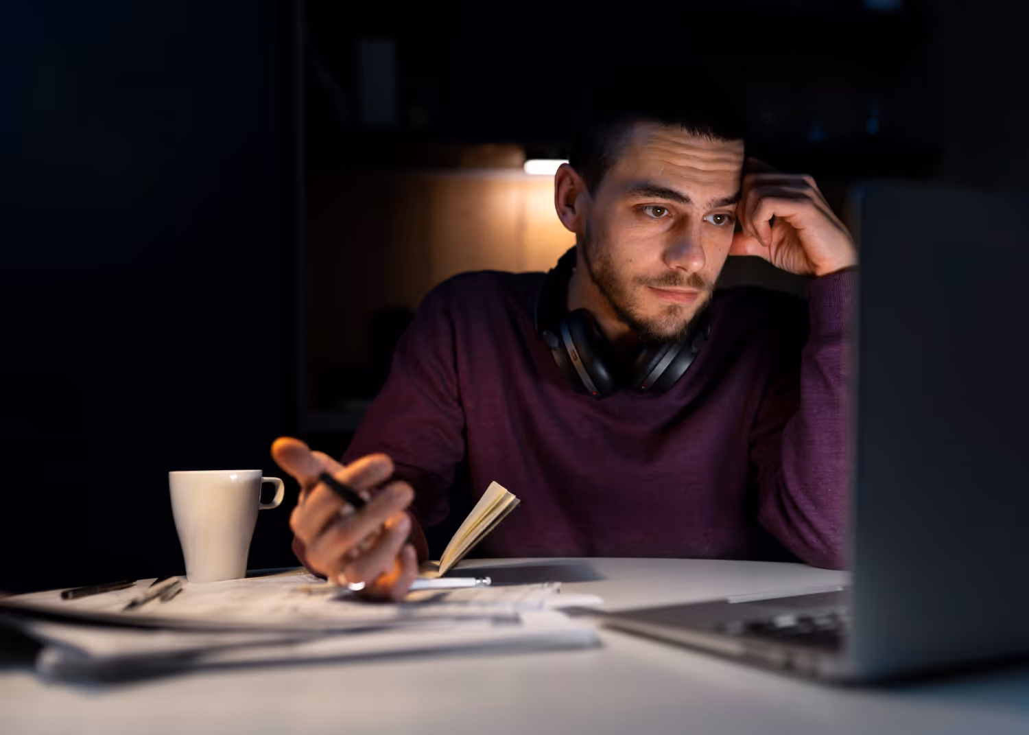 Man in a purple sweater studying with papers, notebook, and laptop at a dimly lit desk.